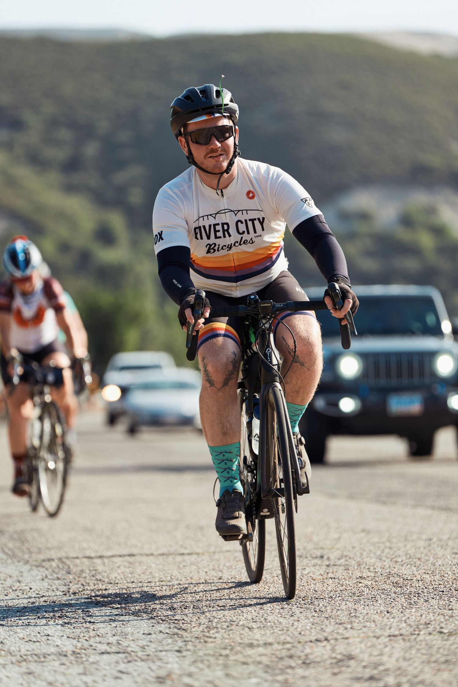 Low angle view of cyclists racing on a gravel road with cars and mountains in the background.
