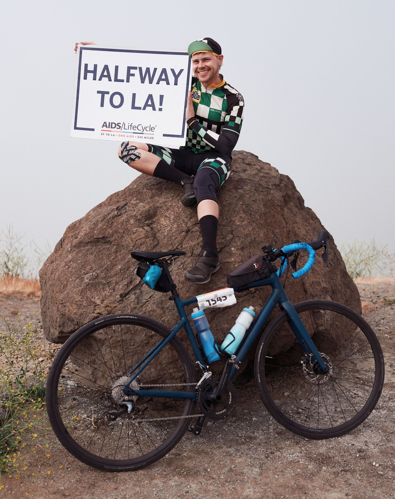 Cyclist resting on a large boulder with a blue gravel bike and 'Halfway to LA' sign in a desert landscape.