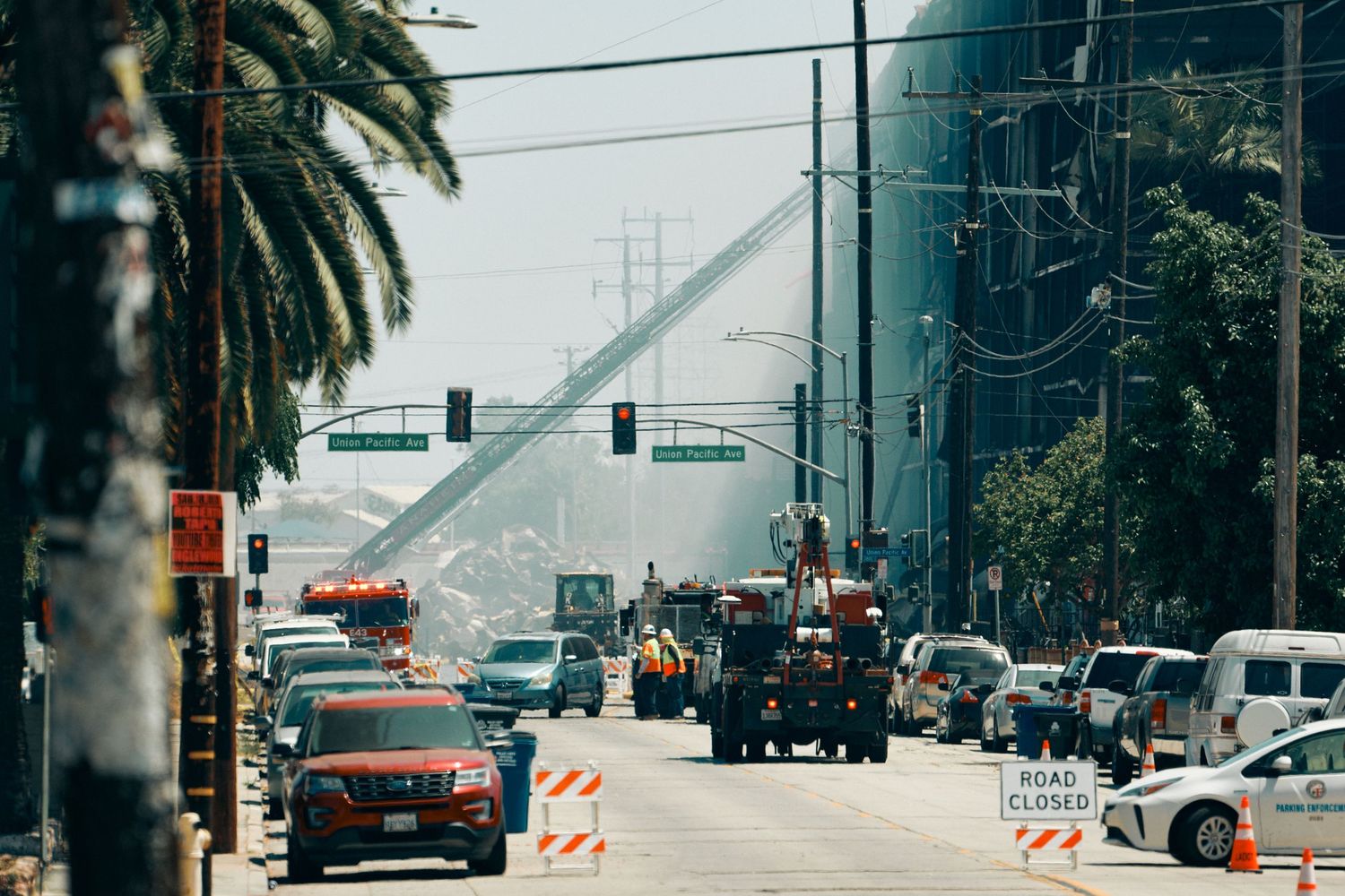 Urban street with traffic, construction vehicles, orange barriers, a Road Closed sign, palm trees, and a large crane in smoky haze.