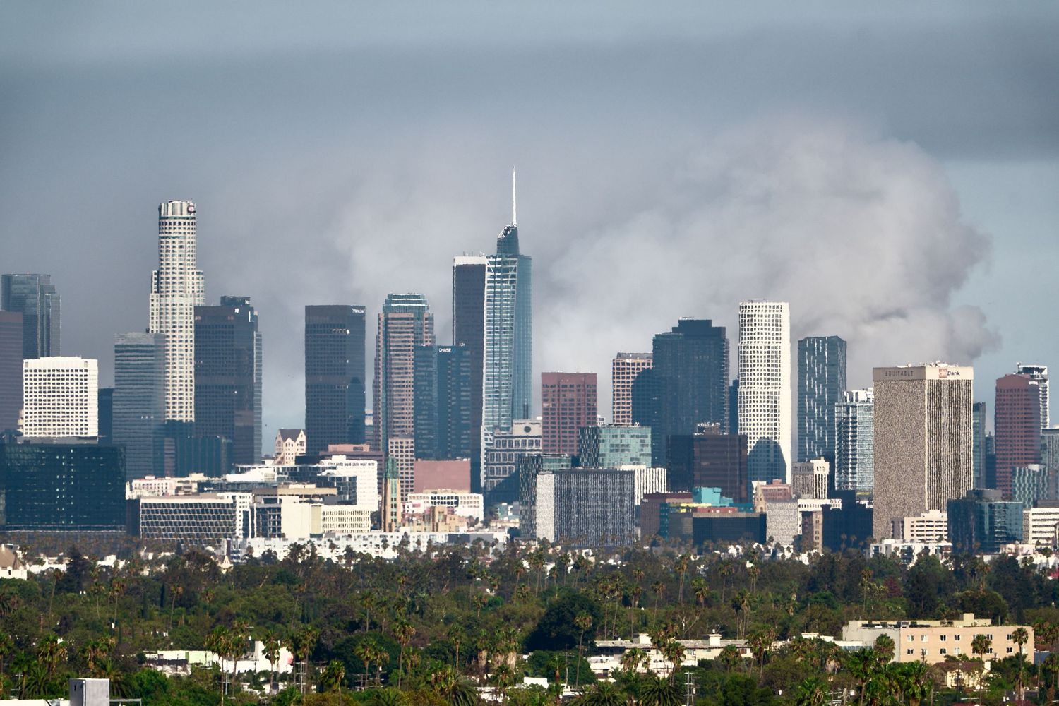 Downtown Los Angeles skyline under a gray sky, with pale smoke rising behind the towers from a distant fire.