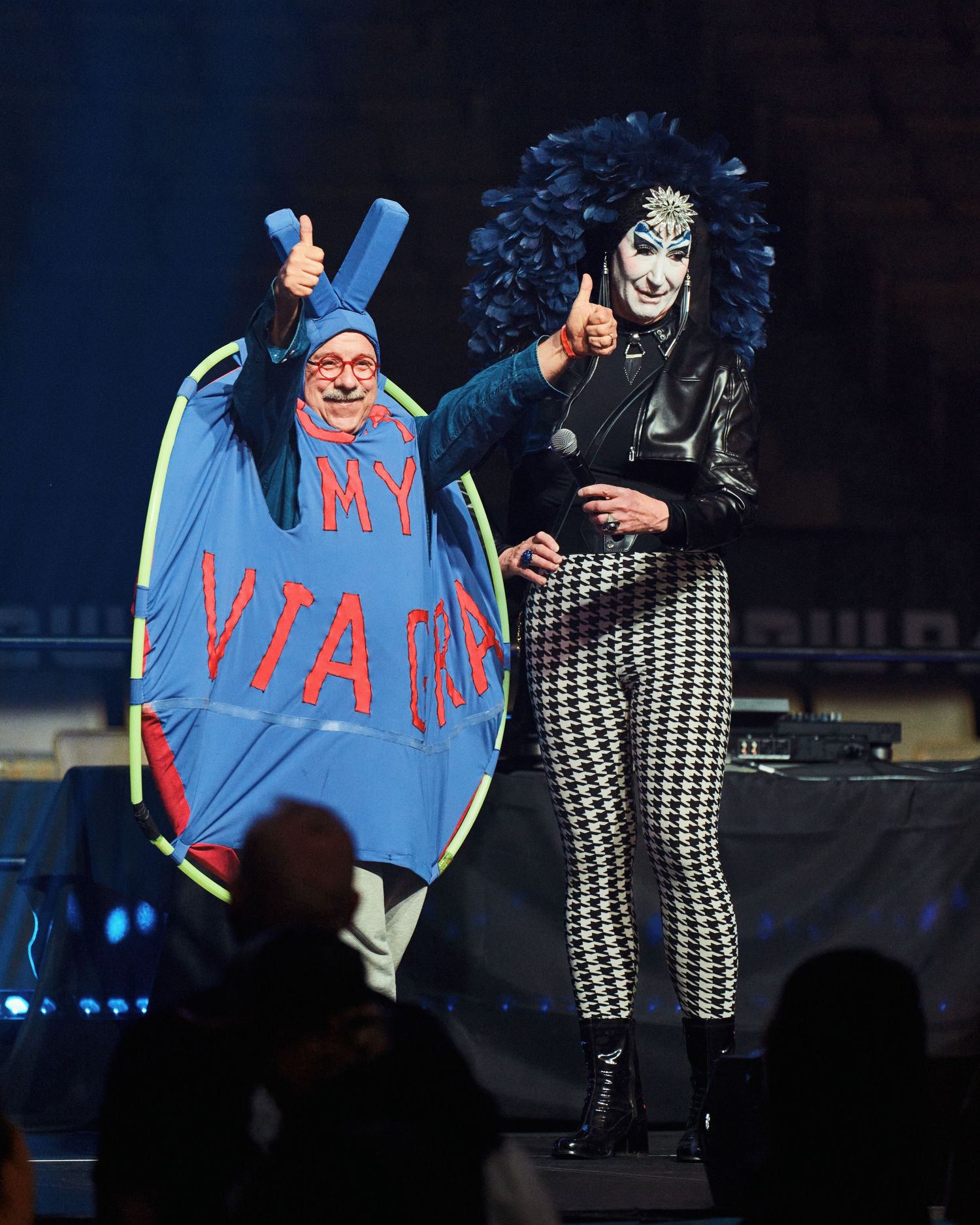 Two performers dancing on stage in contrasting outfits of bright blue and black and white houndstooth pattern costumes.
