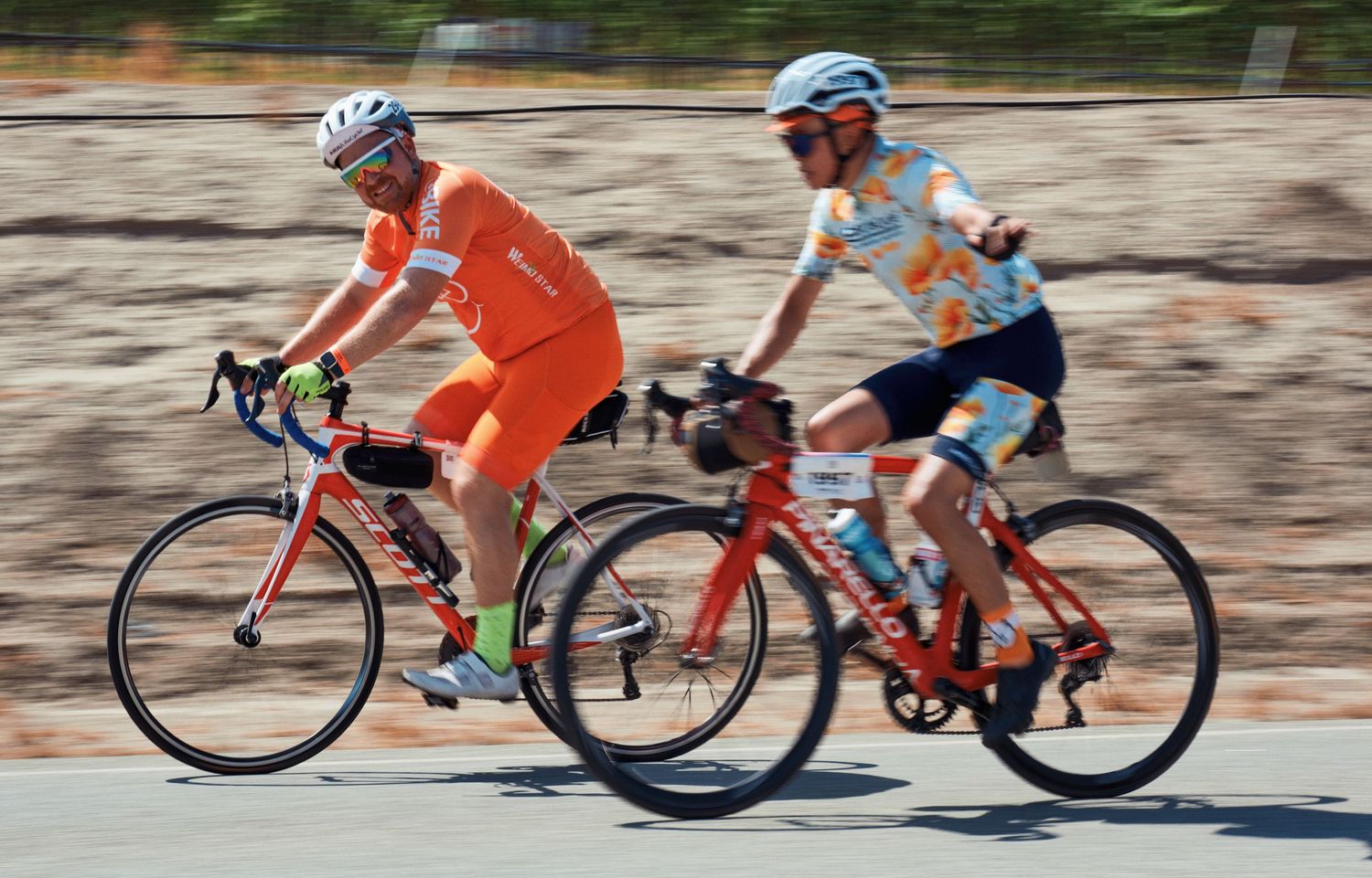 Fast-paced cycling action shot showing riders in orange and blue jerseys racing along paved road.