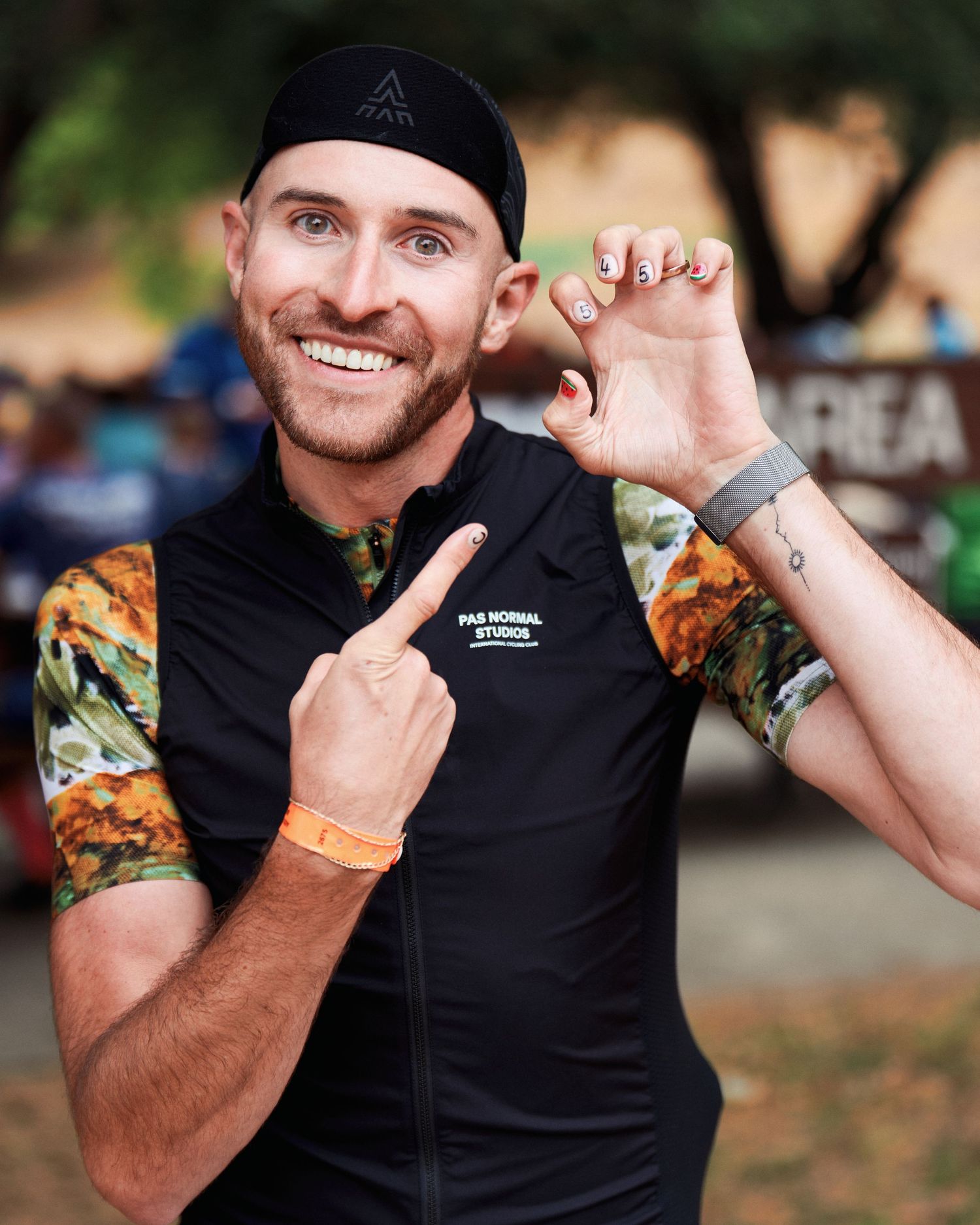 Athletic person in black vest and floral print sleeves smiling and gesturing at outdoor event.