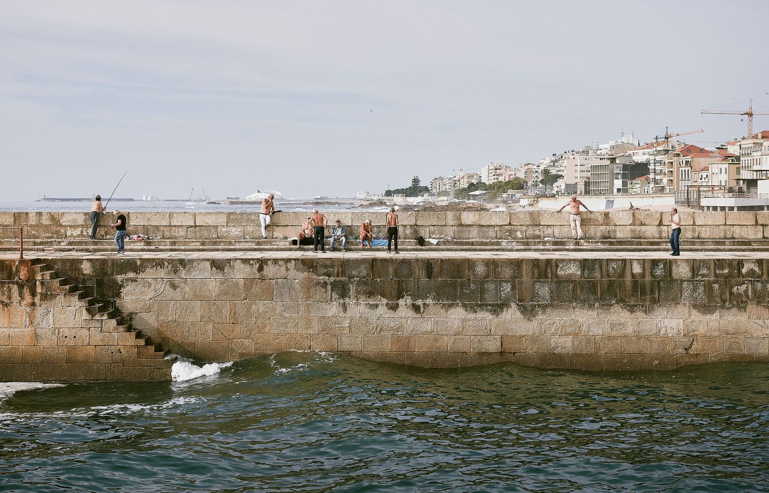 People walking along a stone harbor wall with coastal buildings visible on the hillside in the background.