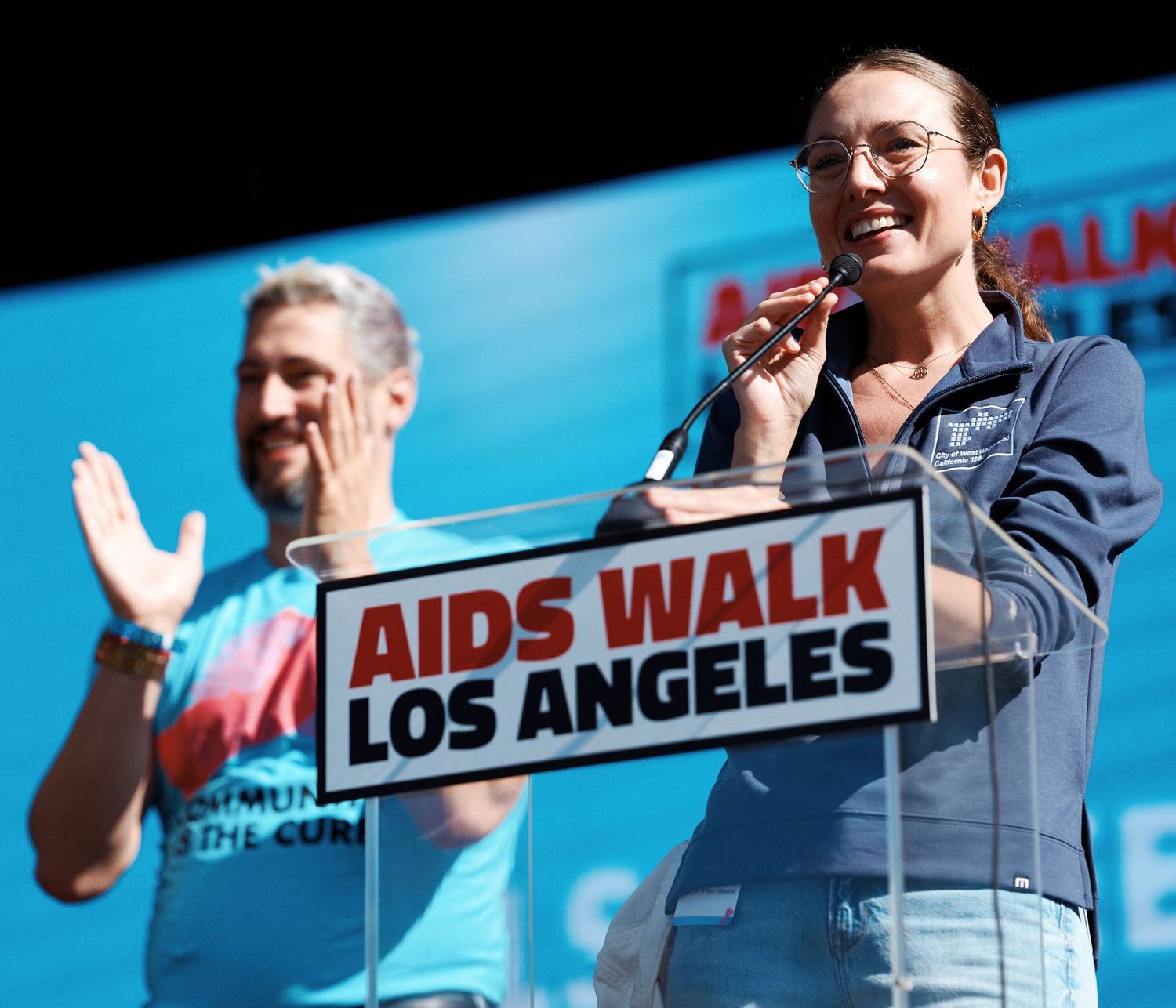 Person in a blue jacket waves to crowd during an AIDS Walk Los Angeles charity event.