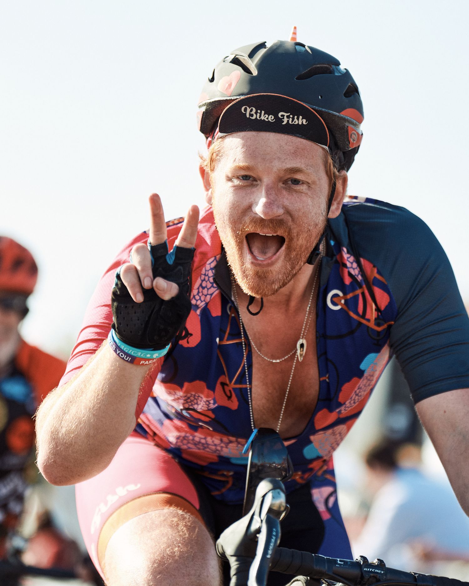 Enthusiastic cyclist wearing a navy and red cycling jersey and helmet gives peace sign while riding outdoors.