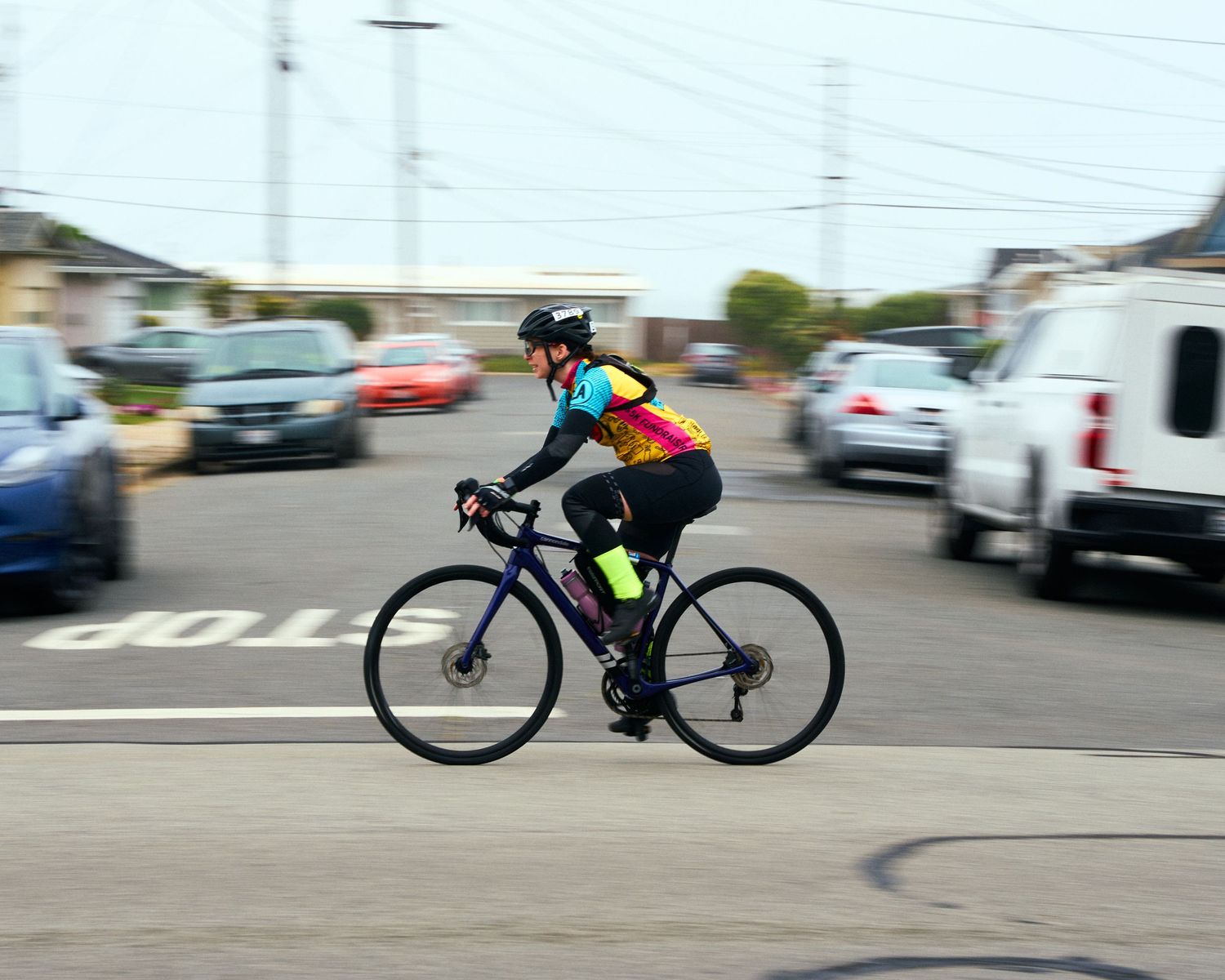 A cyclist in colorful gear rides a black road bike through an intersection with motion blur effect in the background.