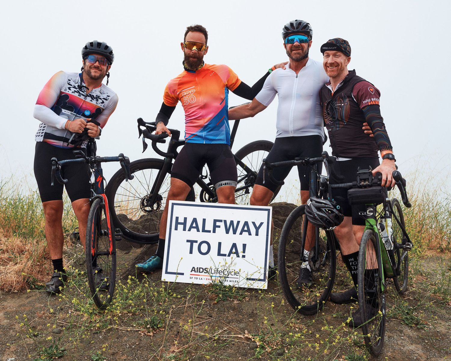 Four cyclists with their bikes pose by a 'Halfway to LA' sign on a hillside trail.