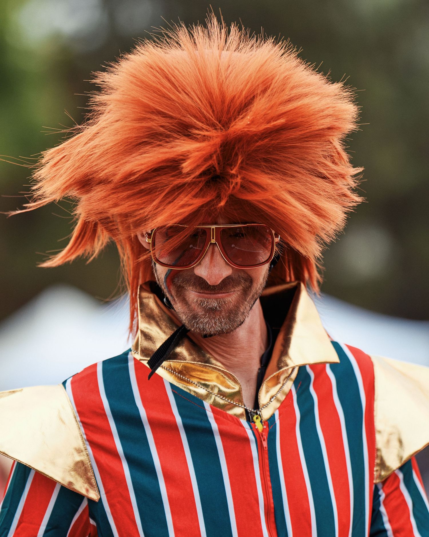 Performer in striped red white and blue outfit with spiky orange hair and glasses performing at an outdoor event.