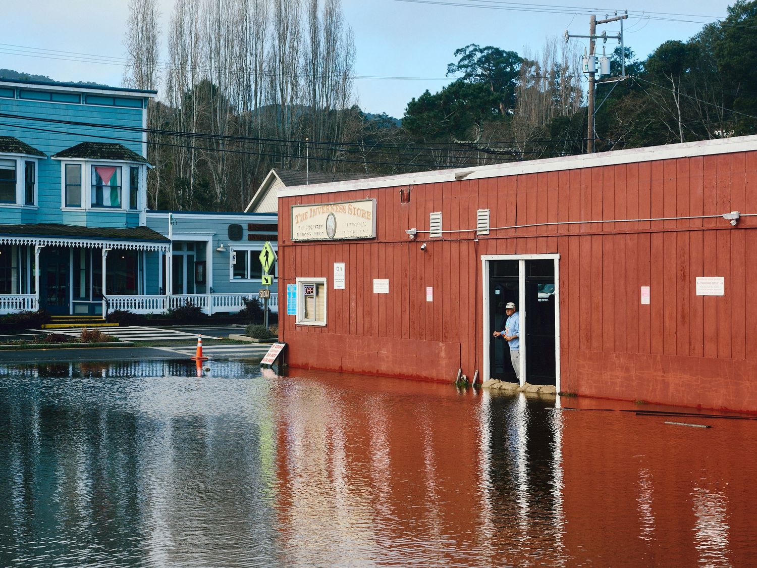 A store owner stands in the doorway of The Inverness Store as floodwaters surround the building in Inverness, Calif., Saturday, Jan. 3, 2026