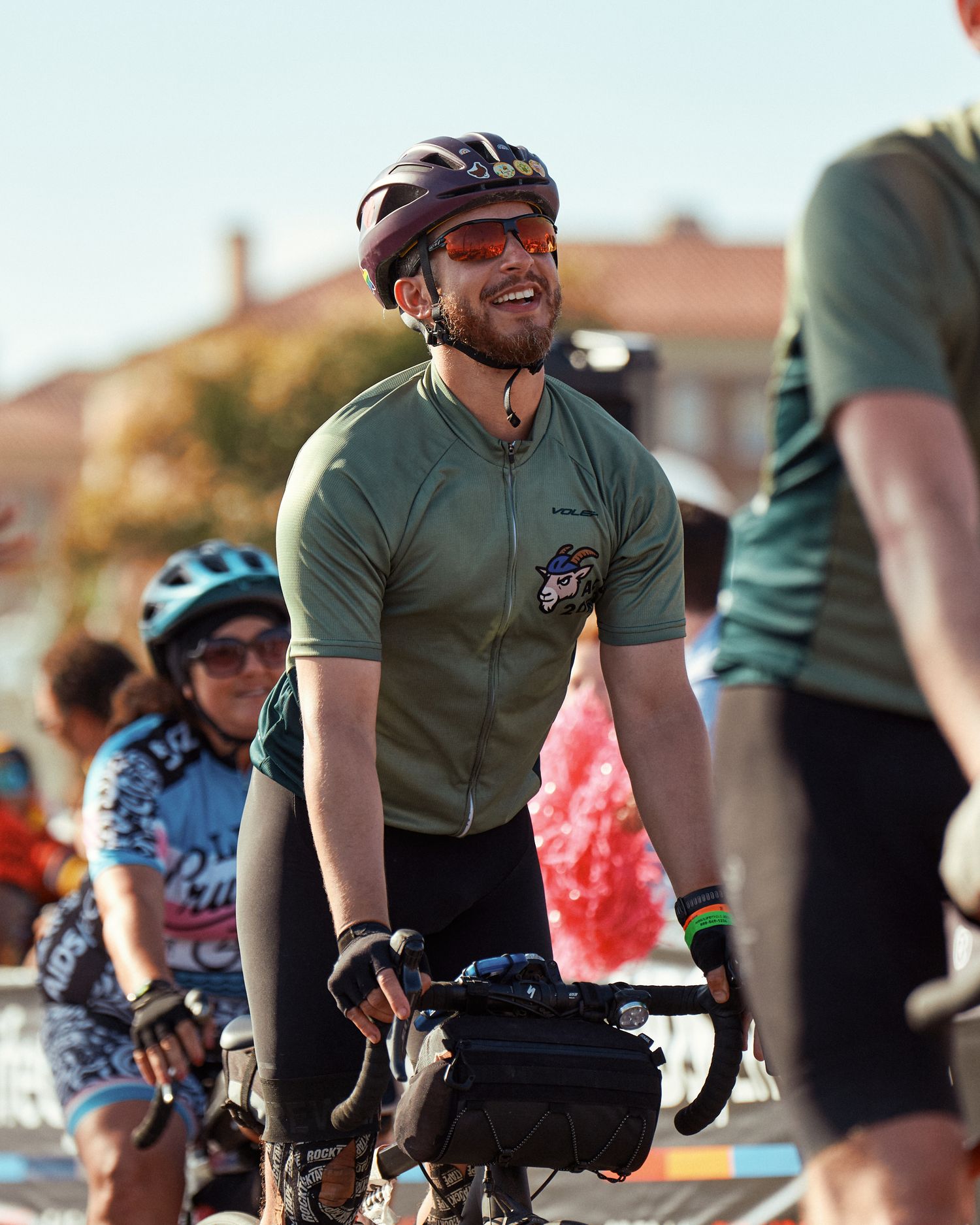 Cyclist in olive green jersey smiling during daytime group ride with other riders visible in background.