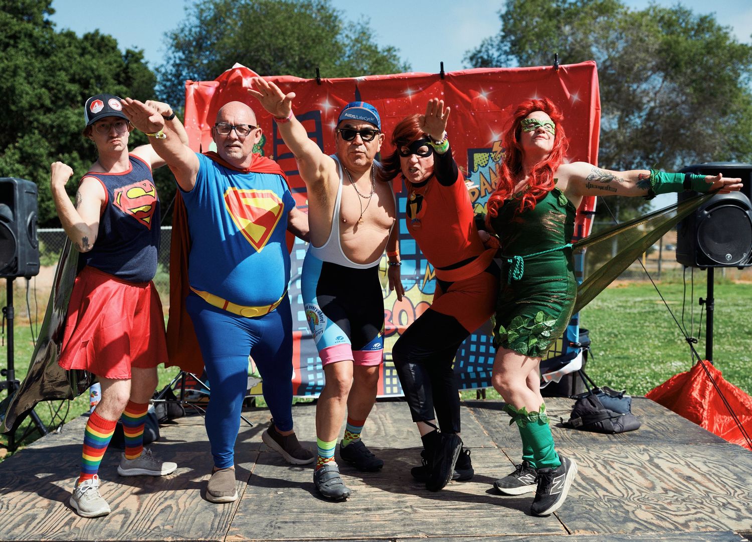 Group of cyclists in colorful costumes including Superman and other superheroes pose together at an outdoor event.
