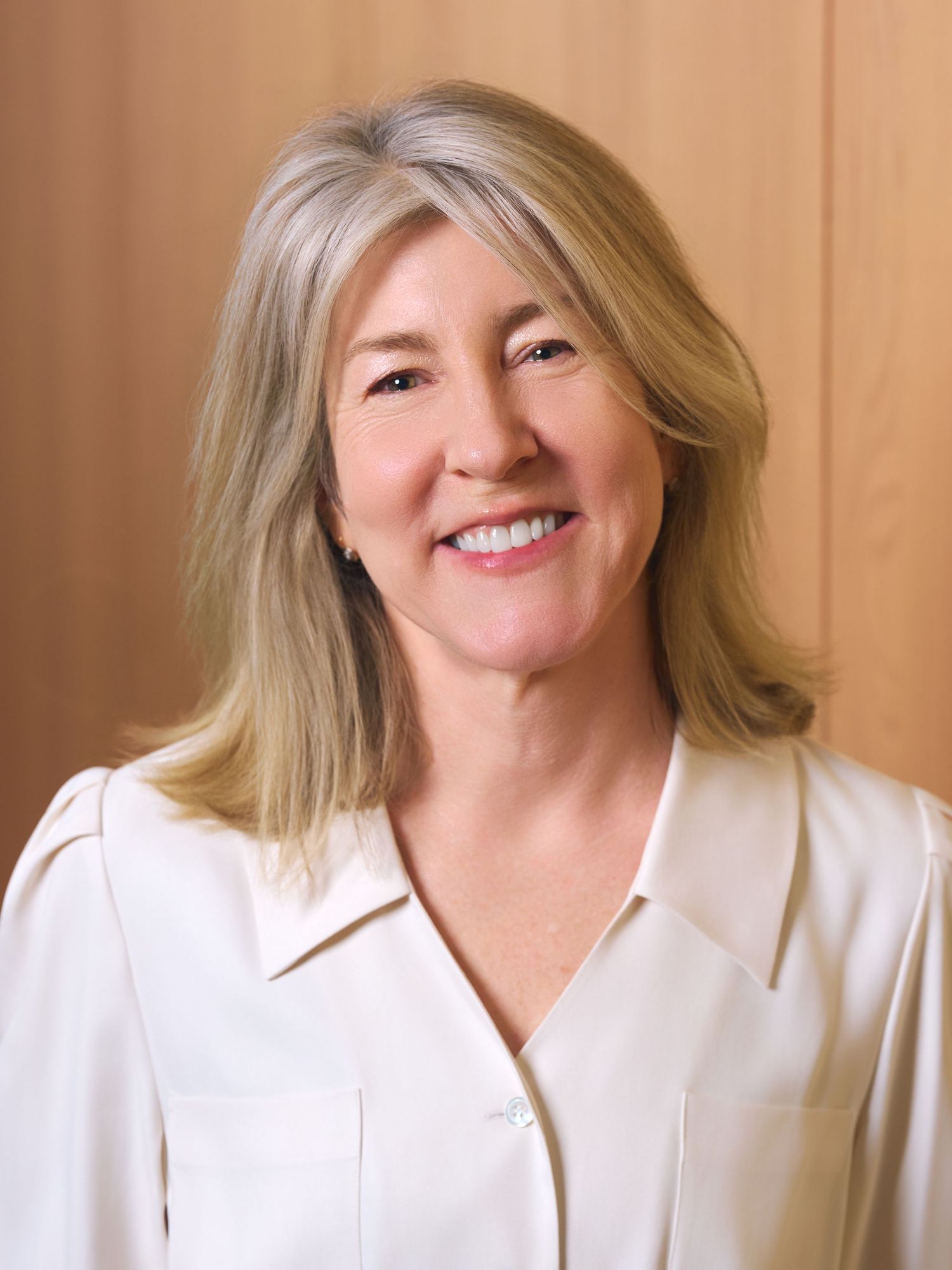 Professional headshot of a smiling middle-aged woman with blonde hair wearing a white blouse against a wood background.