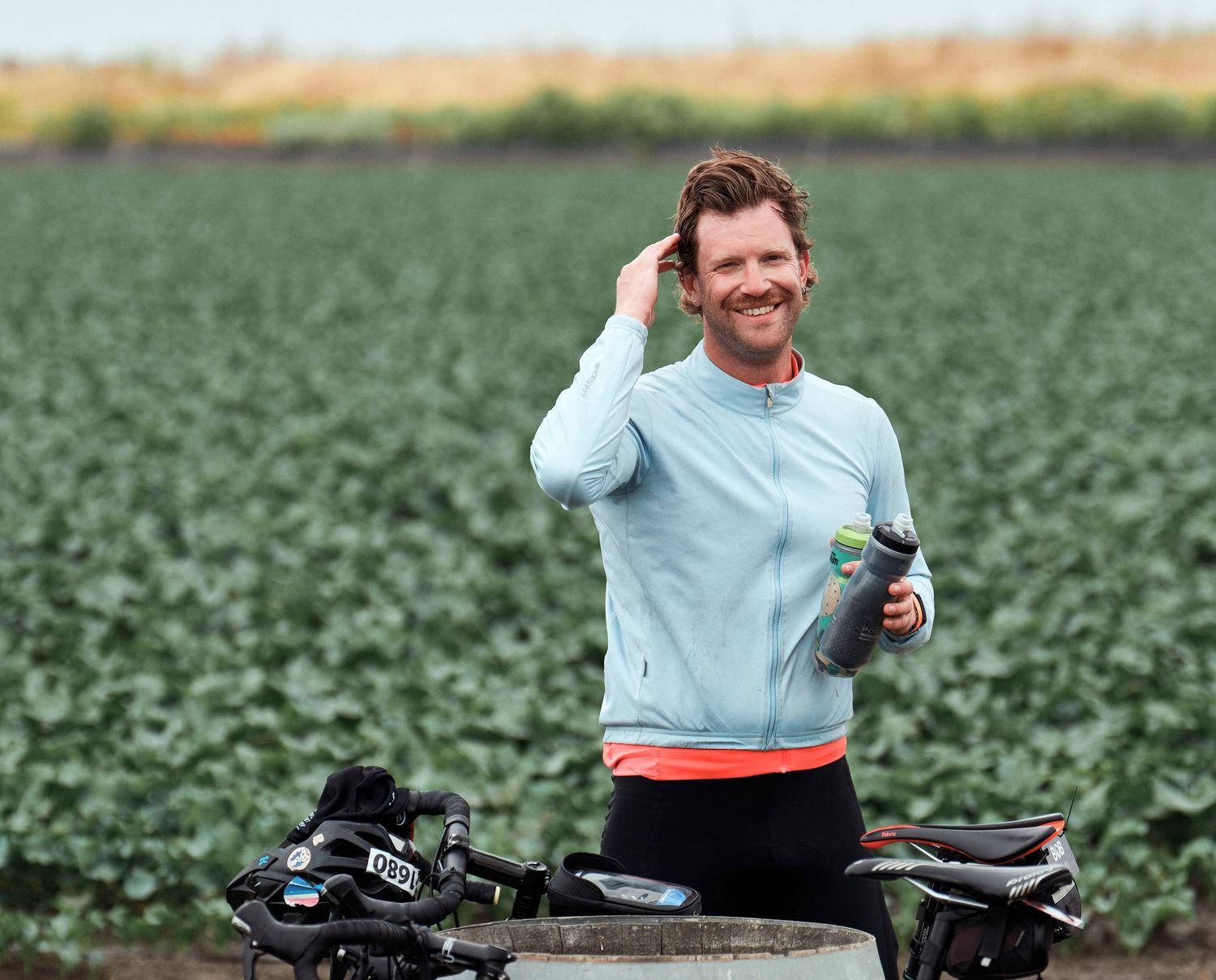 Athletic cyclist in light blue sweater takes a break next to bike while standing in green agricultural field on sunny day.