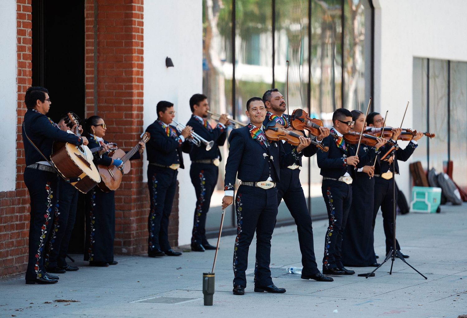 Musicians perform in formal attire with violins along a brick wall during an outdoor concert performance.