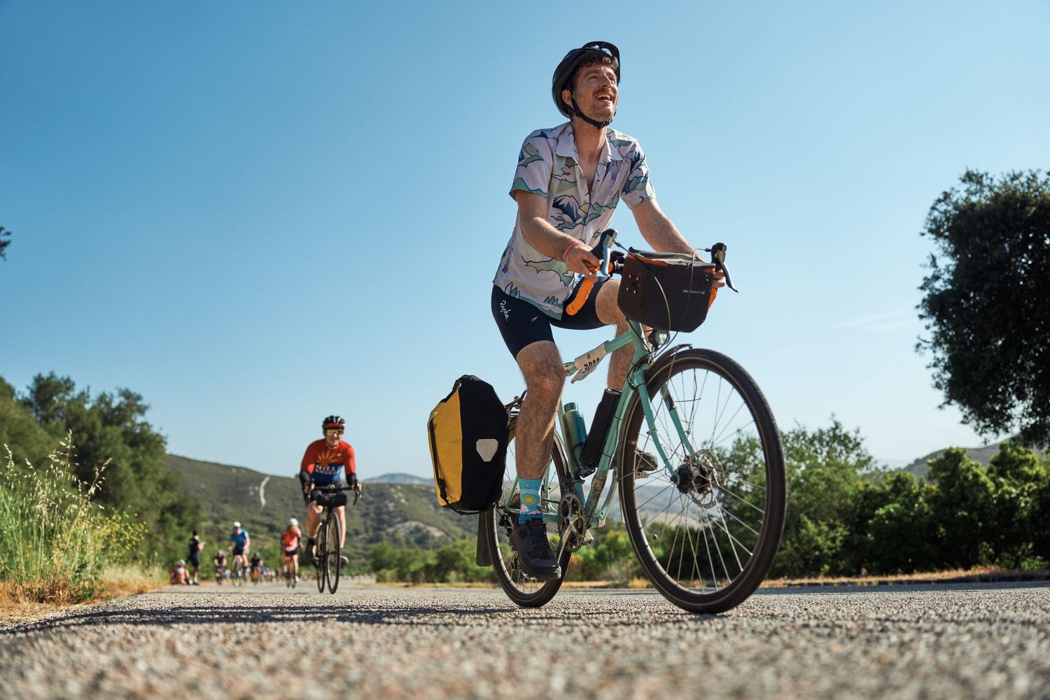Cyclists ride mountain bikes on an outdoor trail and gravel path during daylight hours.