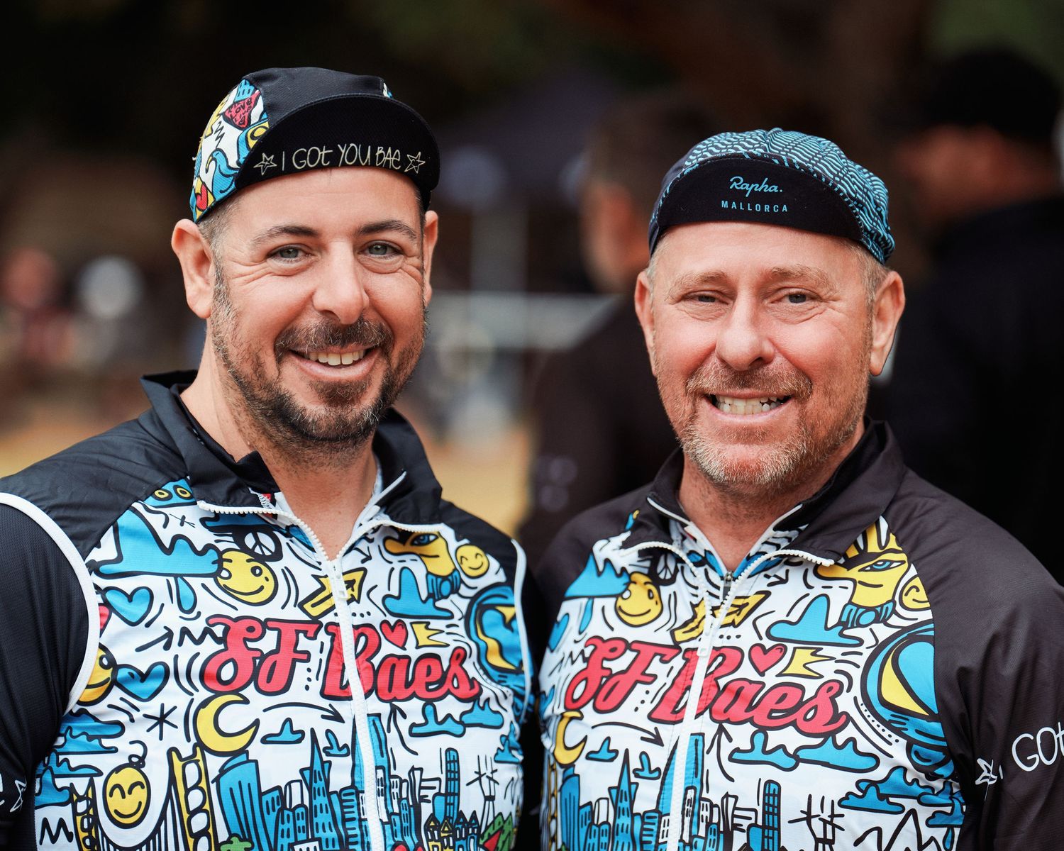 Two people wearing matching colorful graphic print jerseys and baseball caps smile together at outdoor event.