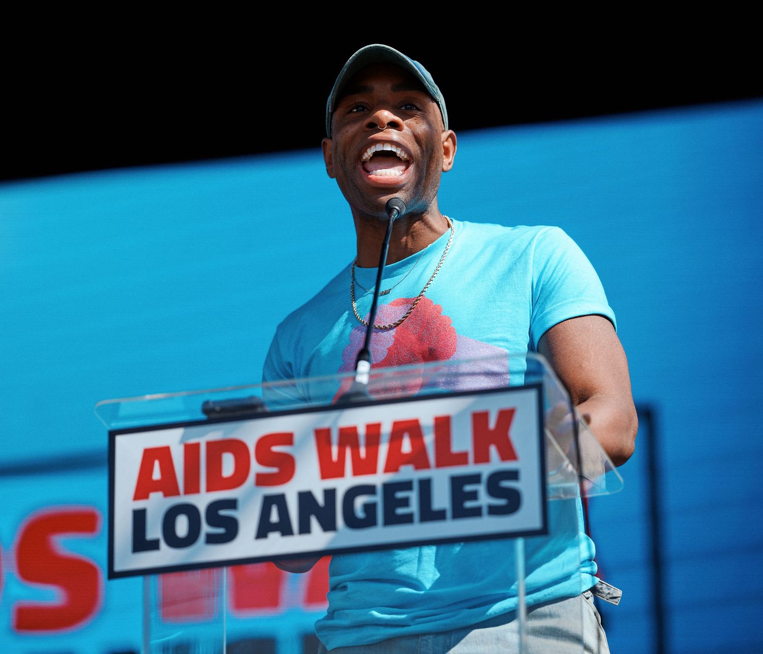 A speaker in a light blue t-shirt smiles while speaking at a podium during the AIDS Walk Los Angeles event.