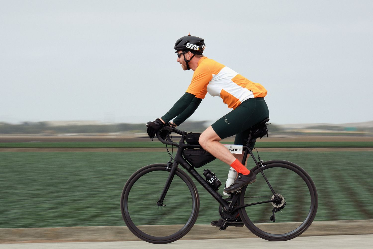A cyclist wearing orange gear rides a road bike through scenic countryside fields and landscape.