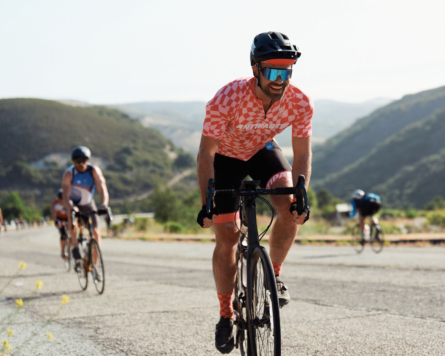 Cyclist in coral jersey rides road bike through mountainous terrain with other riders visible in background.