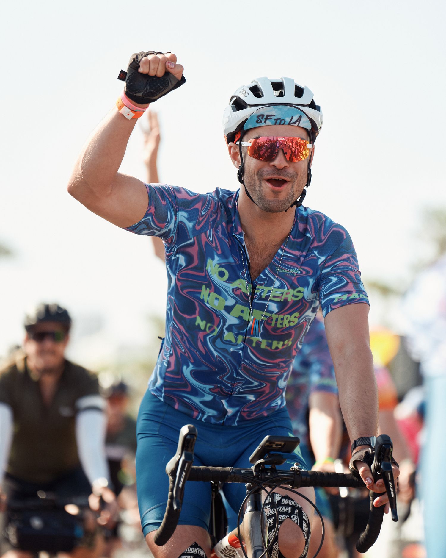 A cyclist in a blue patterned jersey smiles and gestures while riding during an outdoor cycling event or race.