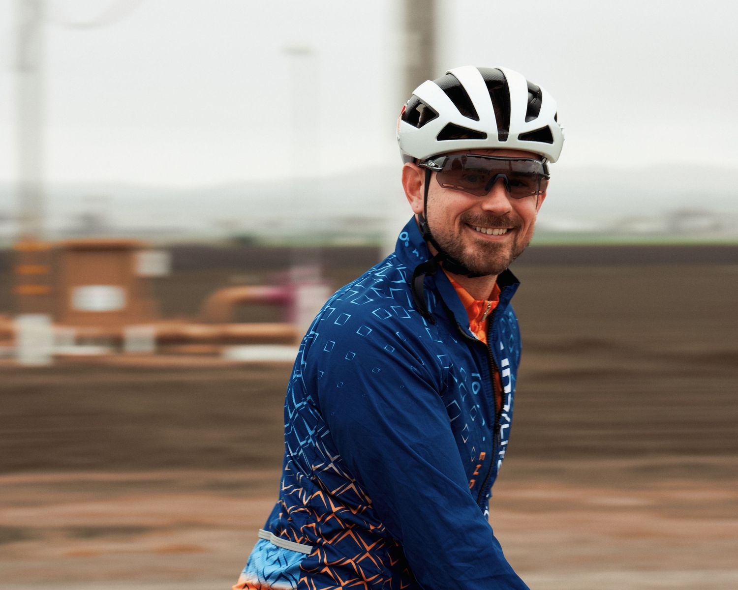 Cyclist in blue patterned jersey and white helmet rides on a cloudy day along rural road.