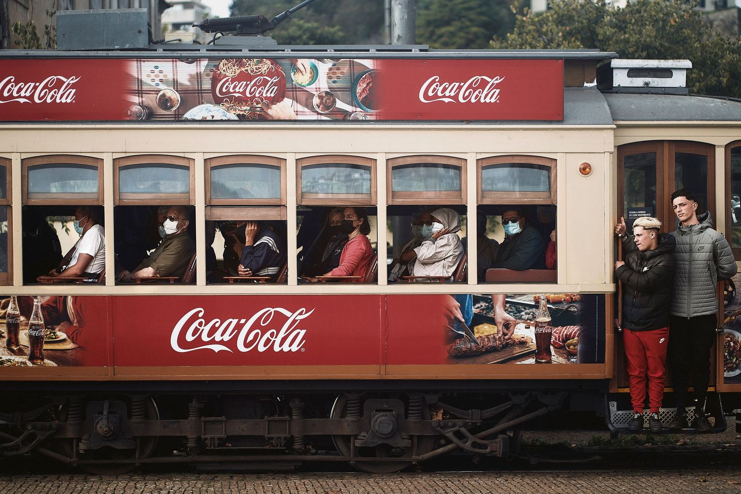A vintage red Coca-Cola themed streetcar filled with passengers standing and sitting in windows during daytime.