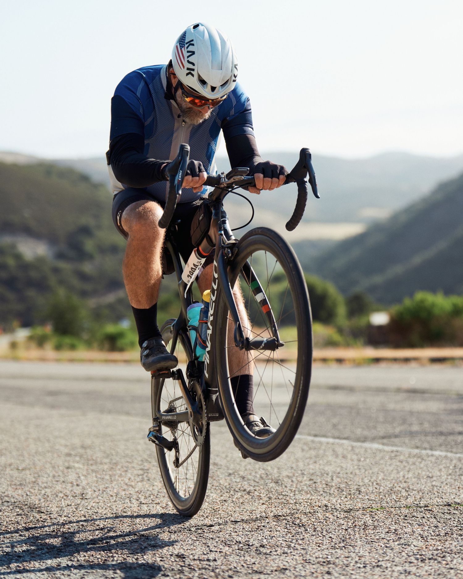 A cyclist performs a wheelie on a road bike against a scenic mountain backdrop on an empty road.