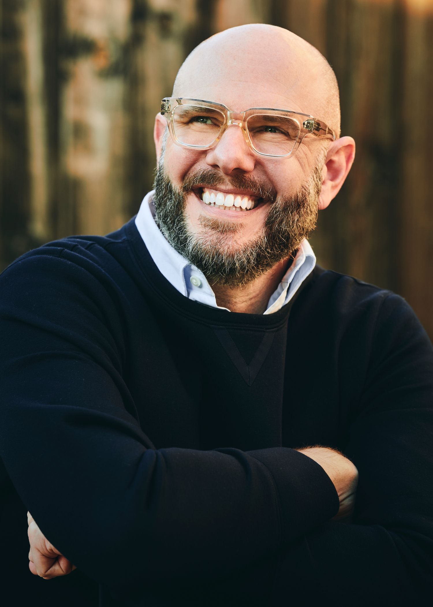 Professional headshot of someone in glasses and a black sweater smiling warmly with arms crossed against a blurred background.