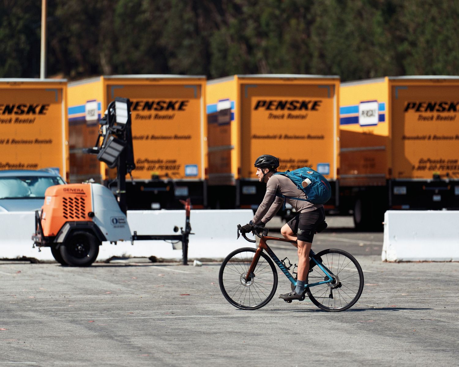 Person riding a bicycle across a parking lot lined with orange Penske rental trucks and barriers.