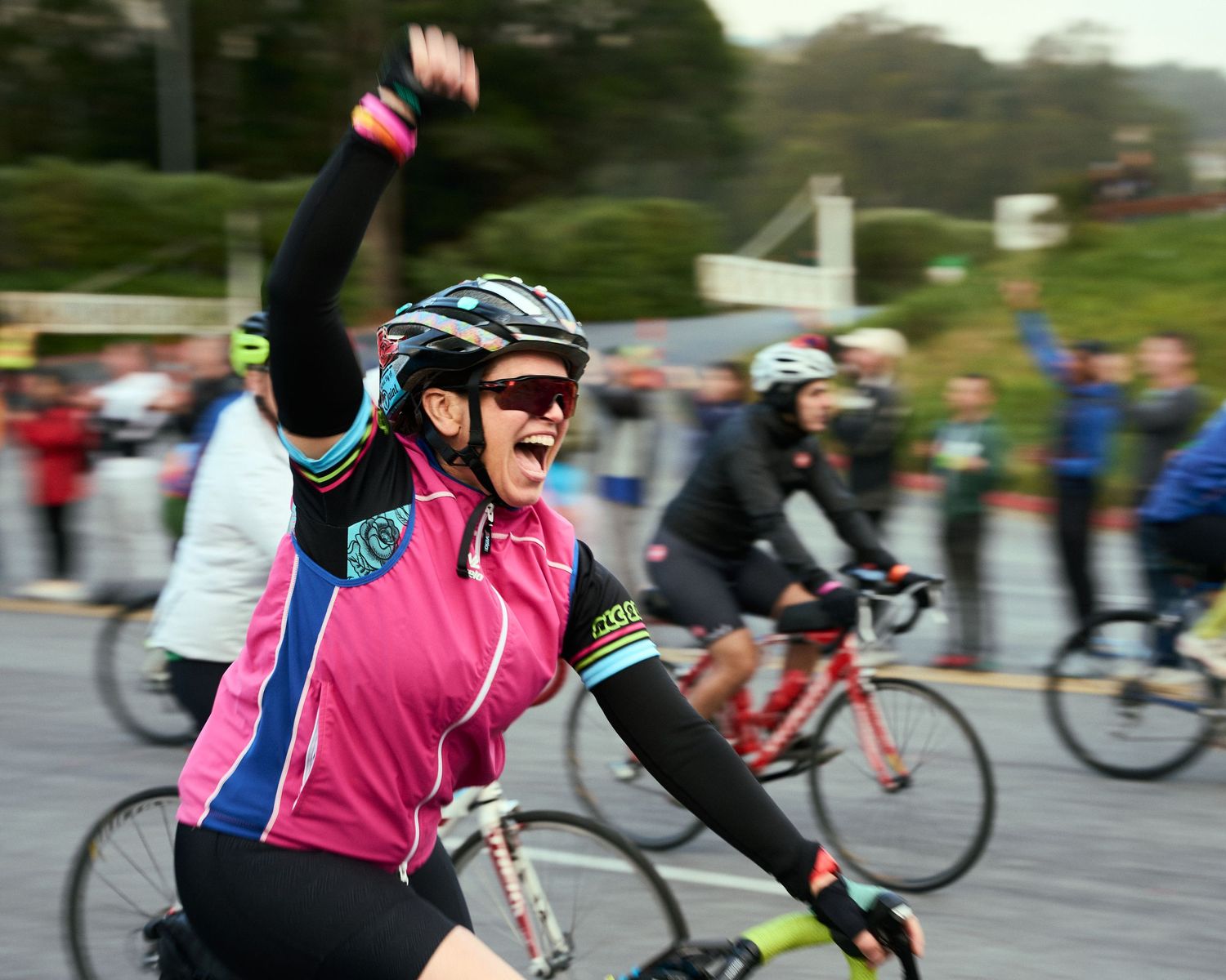 Enthusiastic cyclist in pink jersey raises arm in celebration during group bike ride on cloudy day.