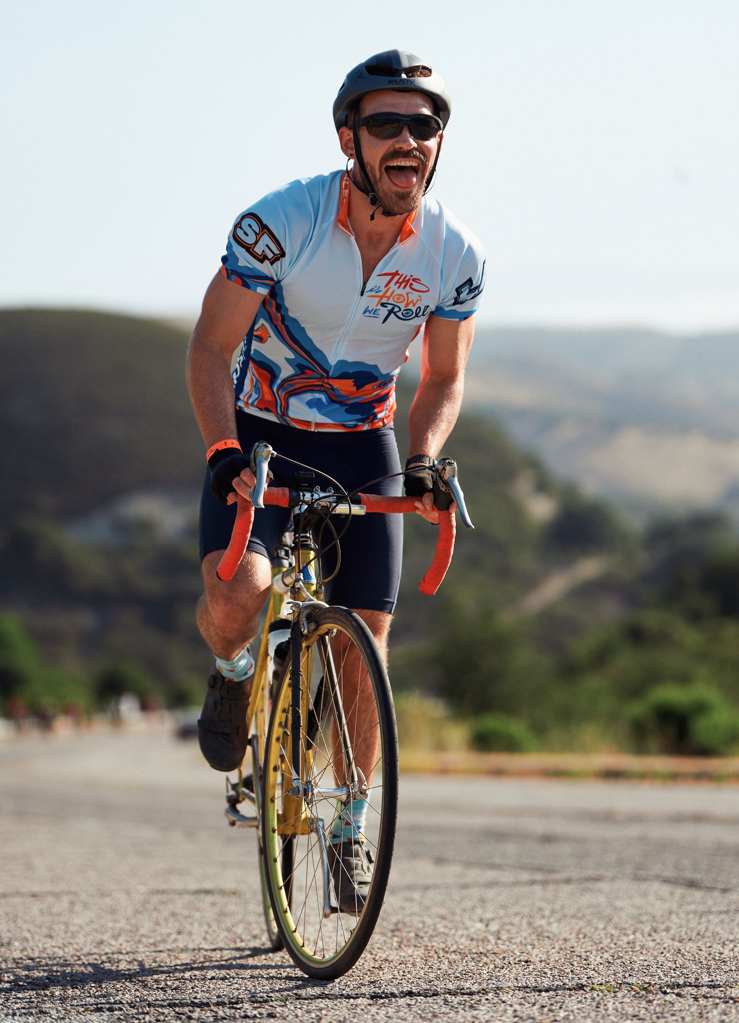 A cyclist in a blue and white jersey rides a road bike on a scenic mountain road on a sunny day.