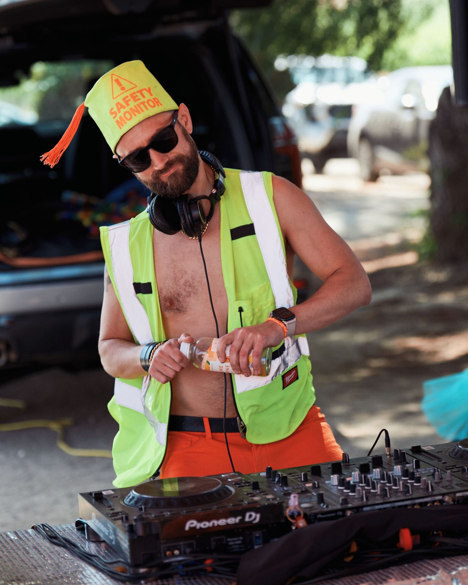DJ in high-vis vest and orange cap performs on Pioneer equipment at outdoor event.