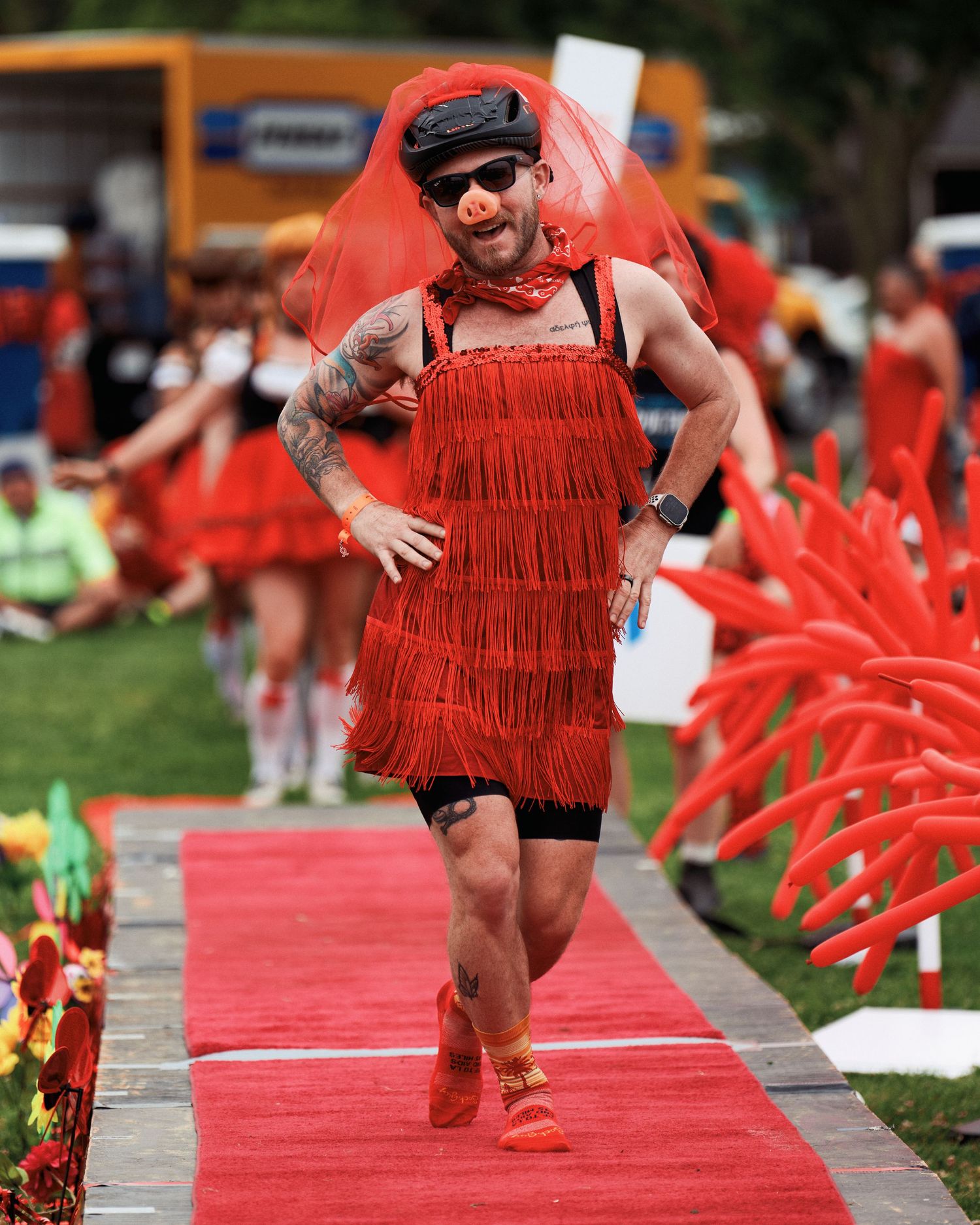 Person in red fringe dress and sneakers walks confidently on a red carpet at an outdoor event.