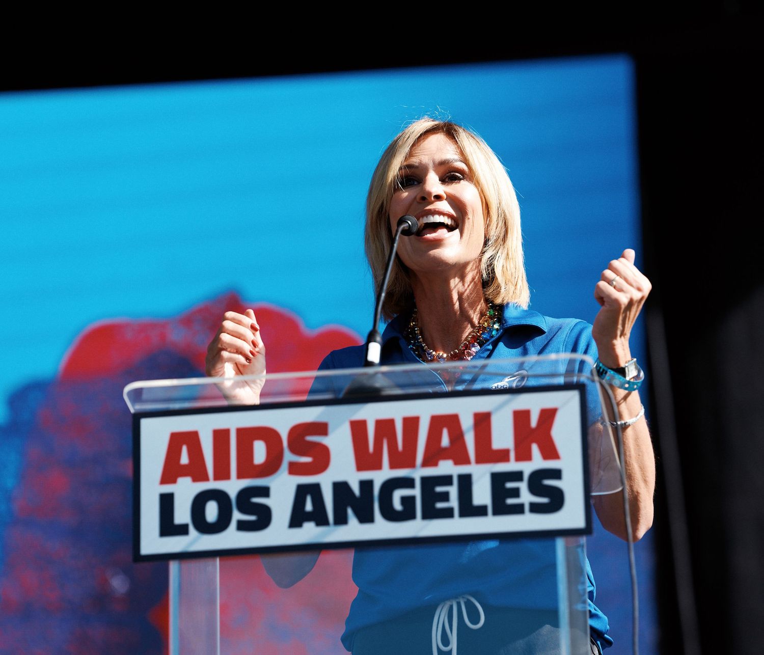 A speaker at an AIDS Walk Los Angeles event stands at a podium against a blue backdrop with signage visible.