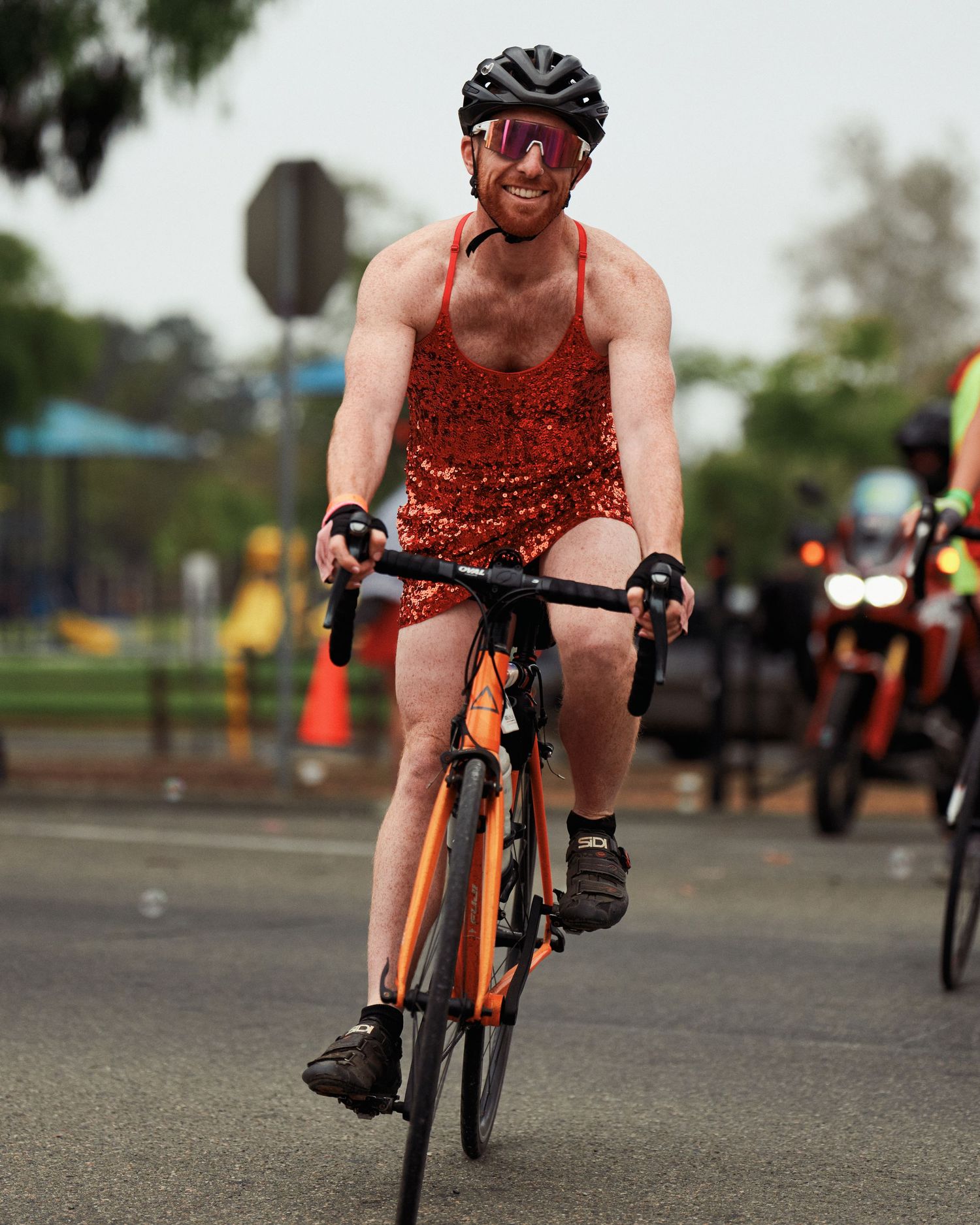 Cyclist in red sparkly outfit and helmet rides road bike during outdoor event.