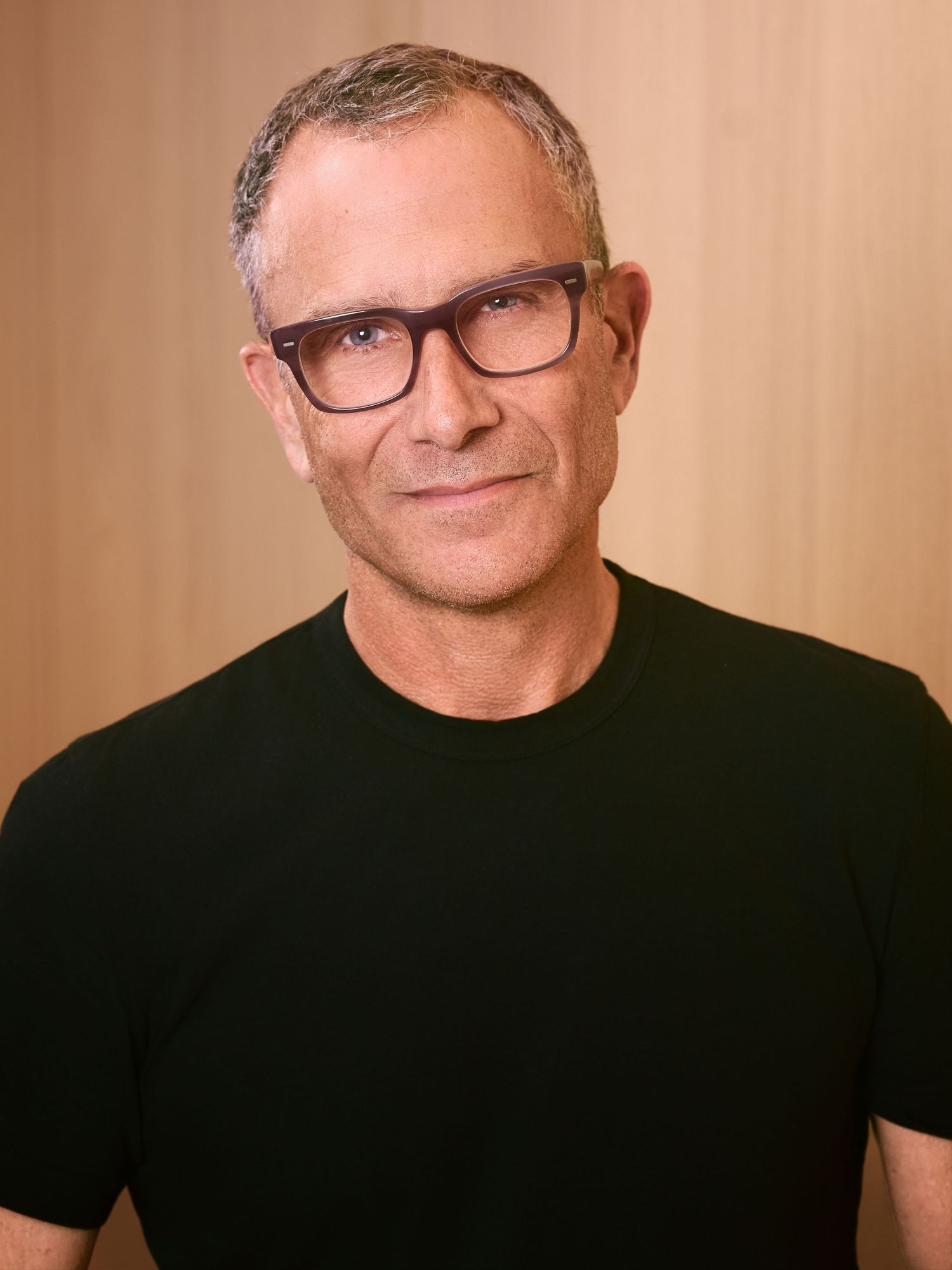 Middle-aged man with short gray hair and dark-rimmed glasses wearing a black t-shirt, posing against a warm wooden background.