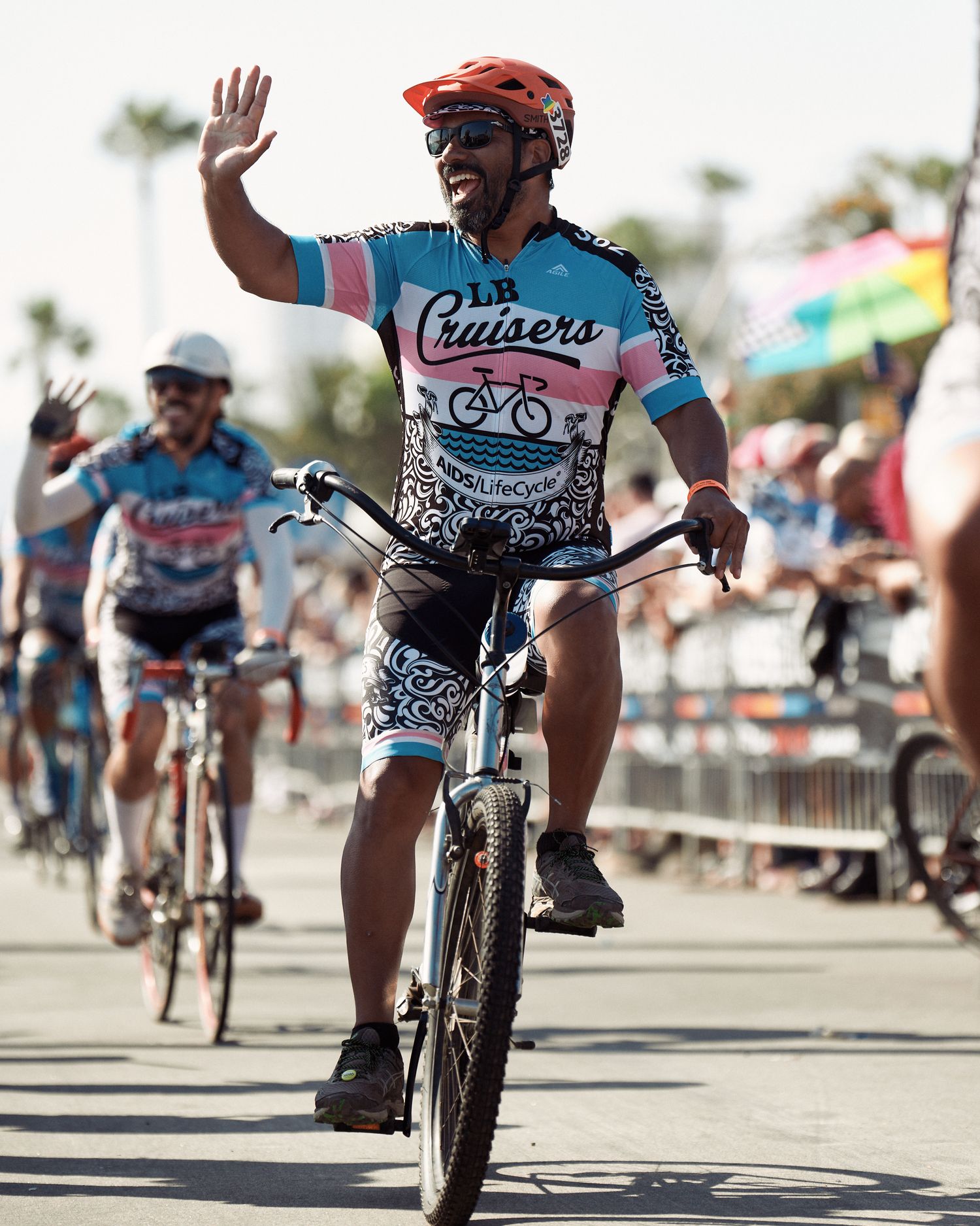 Cyclist in turquoise and white jersey waves while riding mountain bike during outdoor event with palm trees in background.
