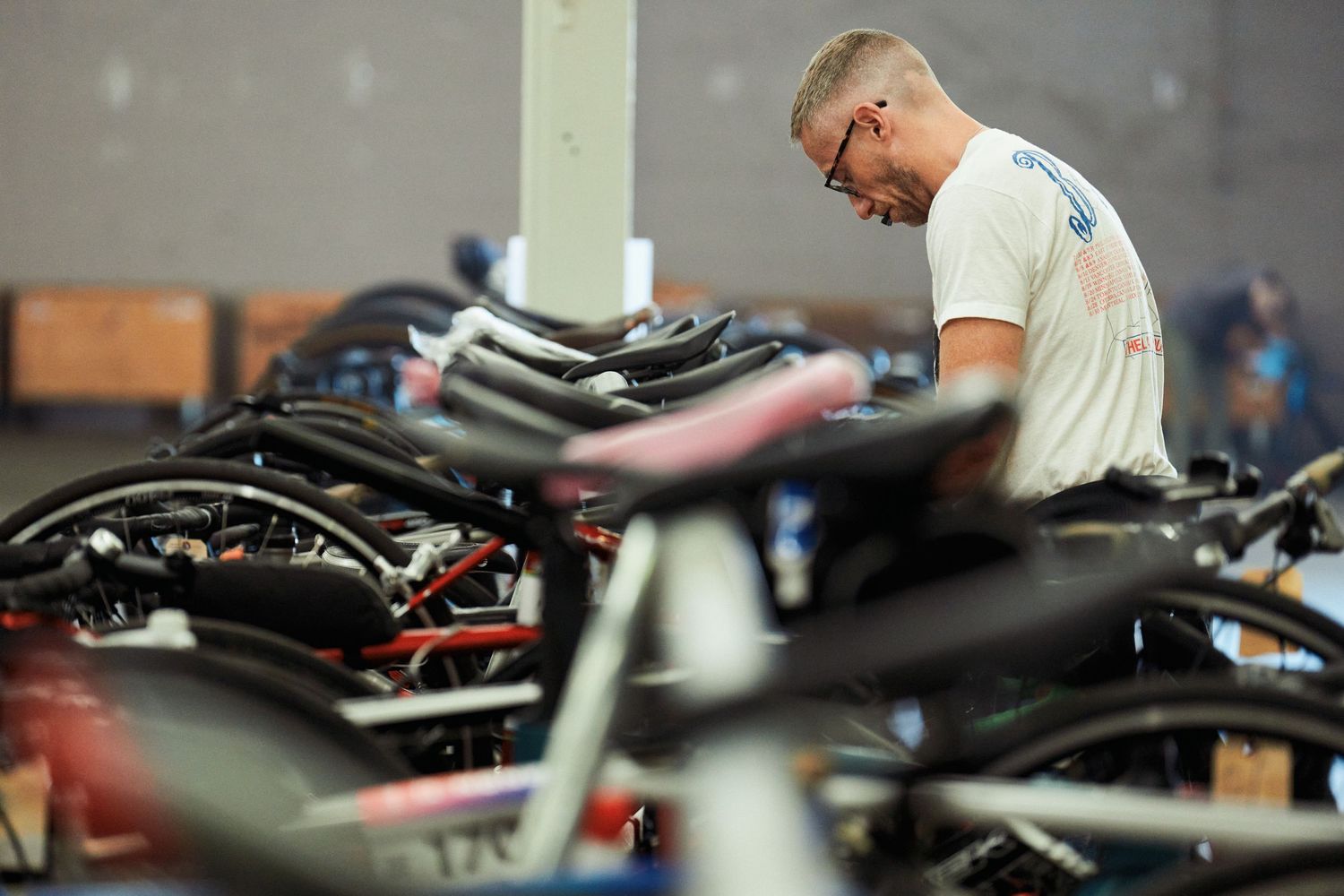 A mechanic in a white t-shirt inspects and works on a collection of parked motorcycles in a garage setting.