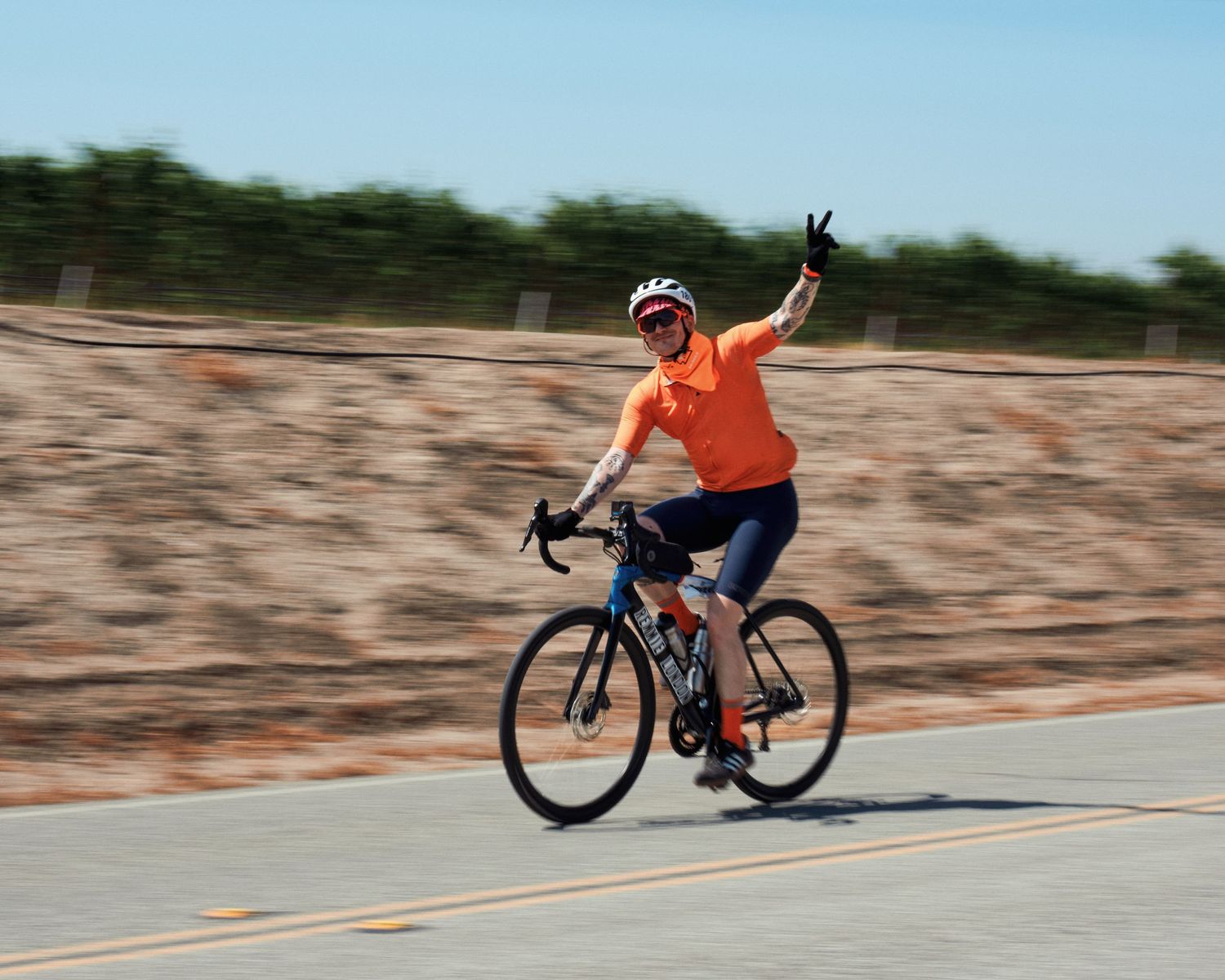 Cyclist in orange safety vest raises arm victoriously while riding on paved road near rocky terrain.
