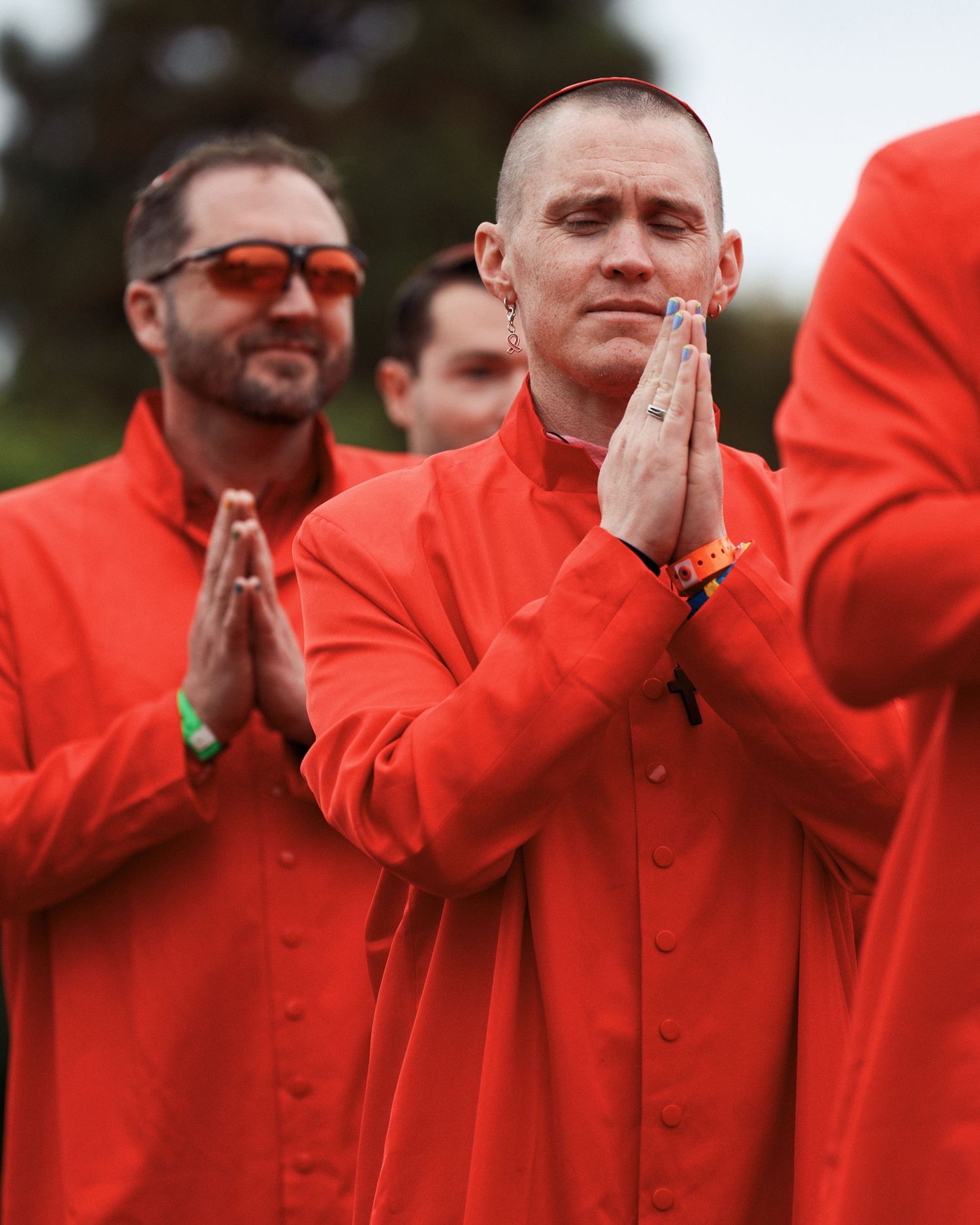Two figures in bright red outfits stand together with prayer-like hand gestures outdoors.