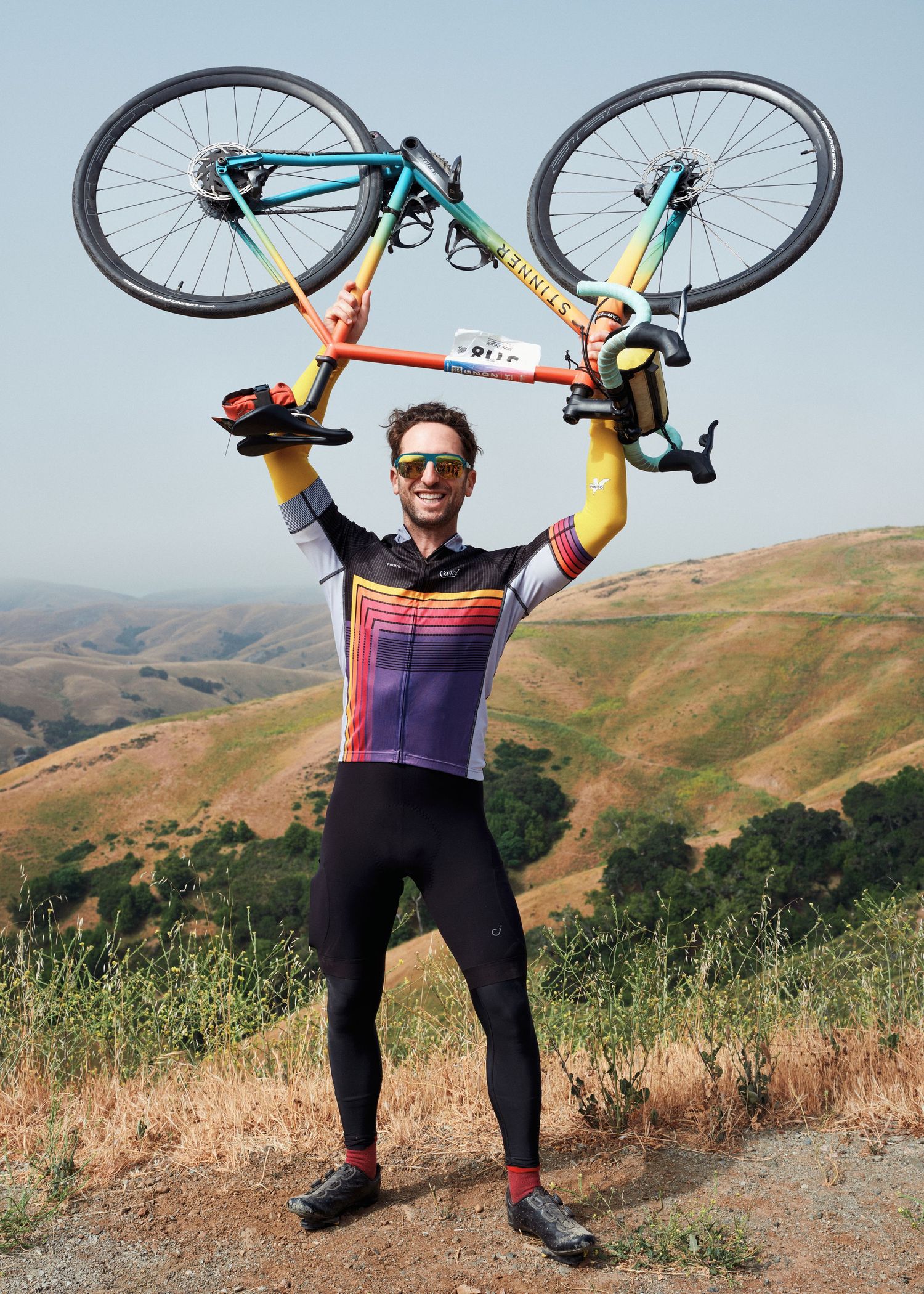 Cyclist in colorful jersey lifts mountain bike overhead triumphantly against scenic rolling hills background.