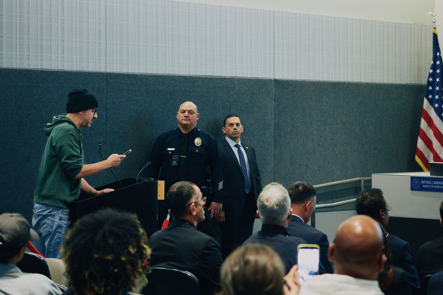 Police officer stands at podium during public meeting with audience and American flag in background.