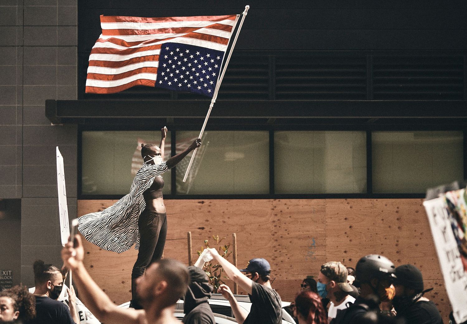 Person waves American flag at a large outdoor protest rally with crowd holding signs in an urban setting.