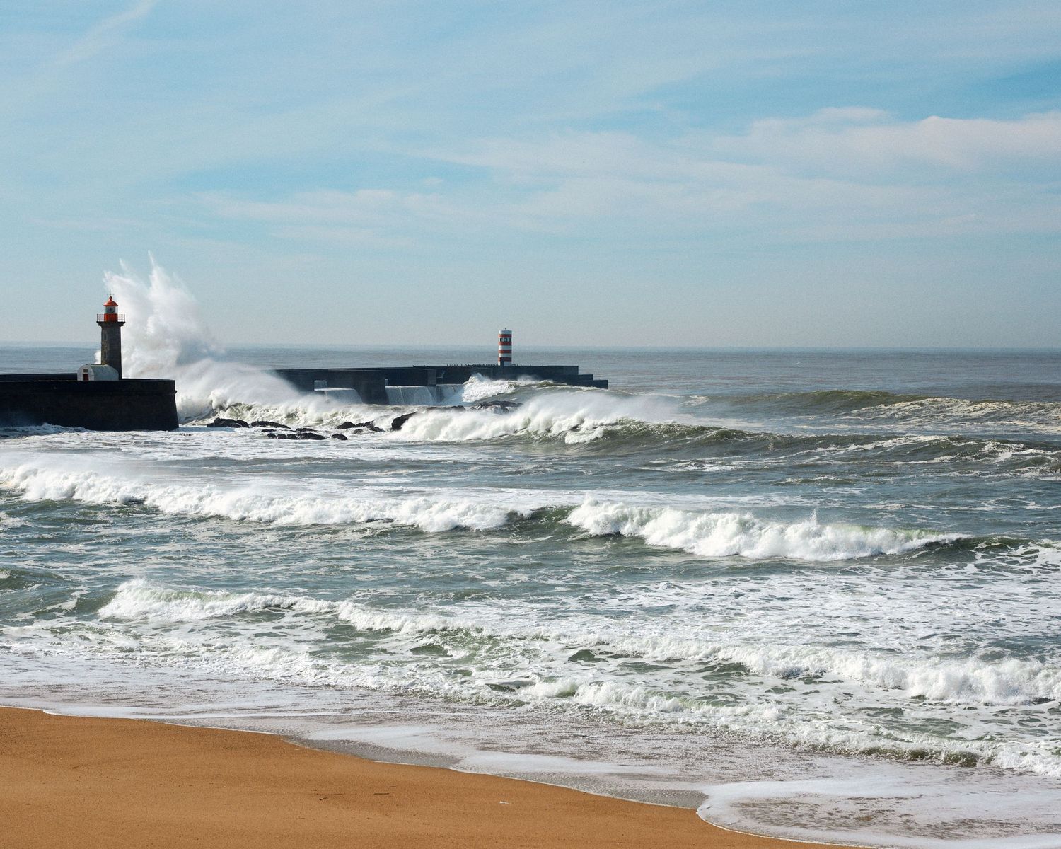 Dramatic waves crash against a lighthouse and rocky coastline during stormy weather at sea.