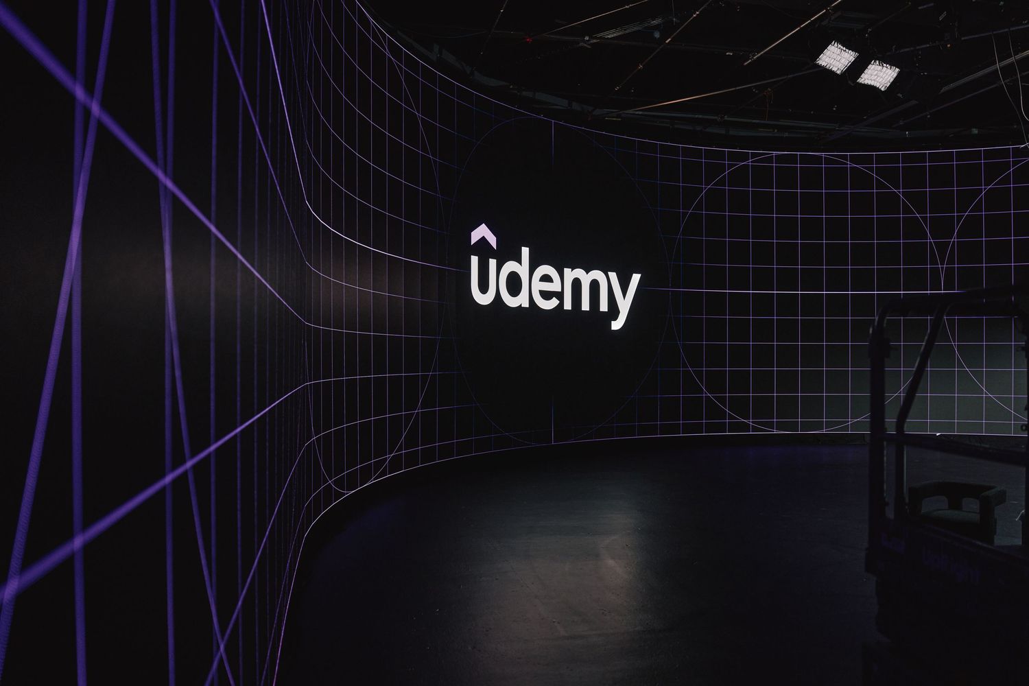 Udemy logo illuminated in dark studio space with geometric wire grid patterns in background.