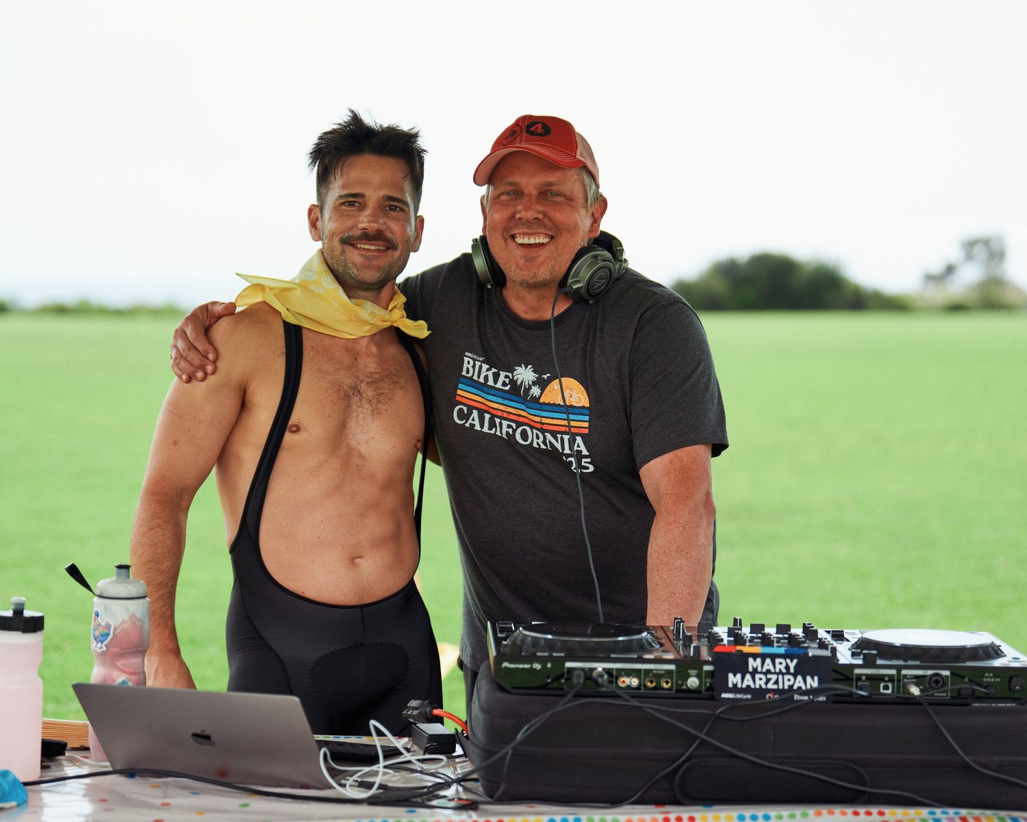 Two DJs performing at outdoor event with equipment and mountains in background.