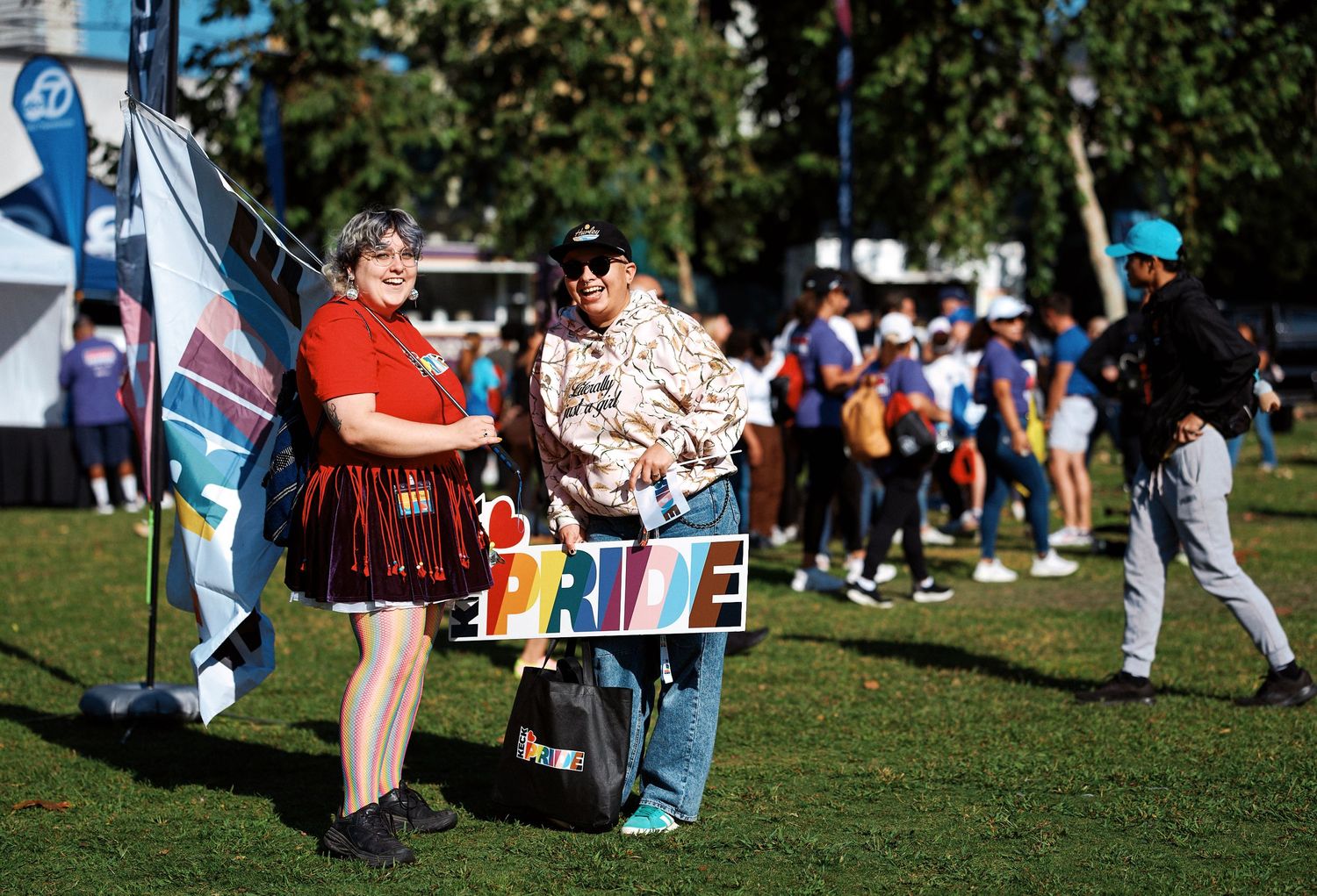 People gather at an outdoor festival with flags and banners on a sunny day in a grassy park area.