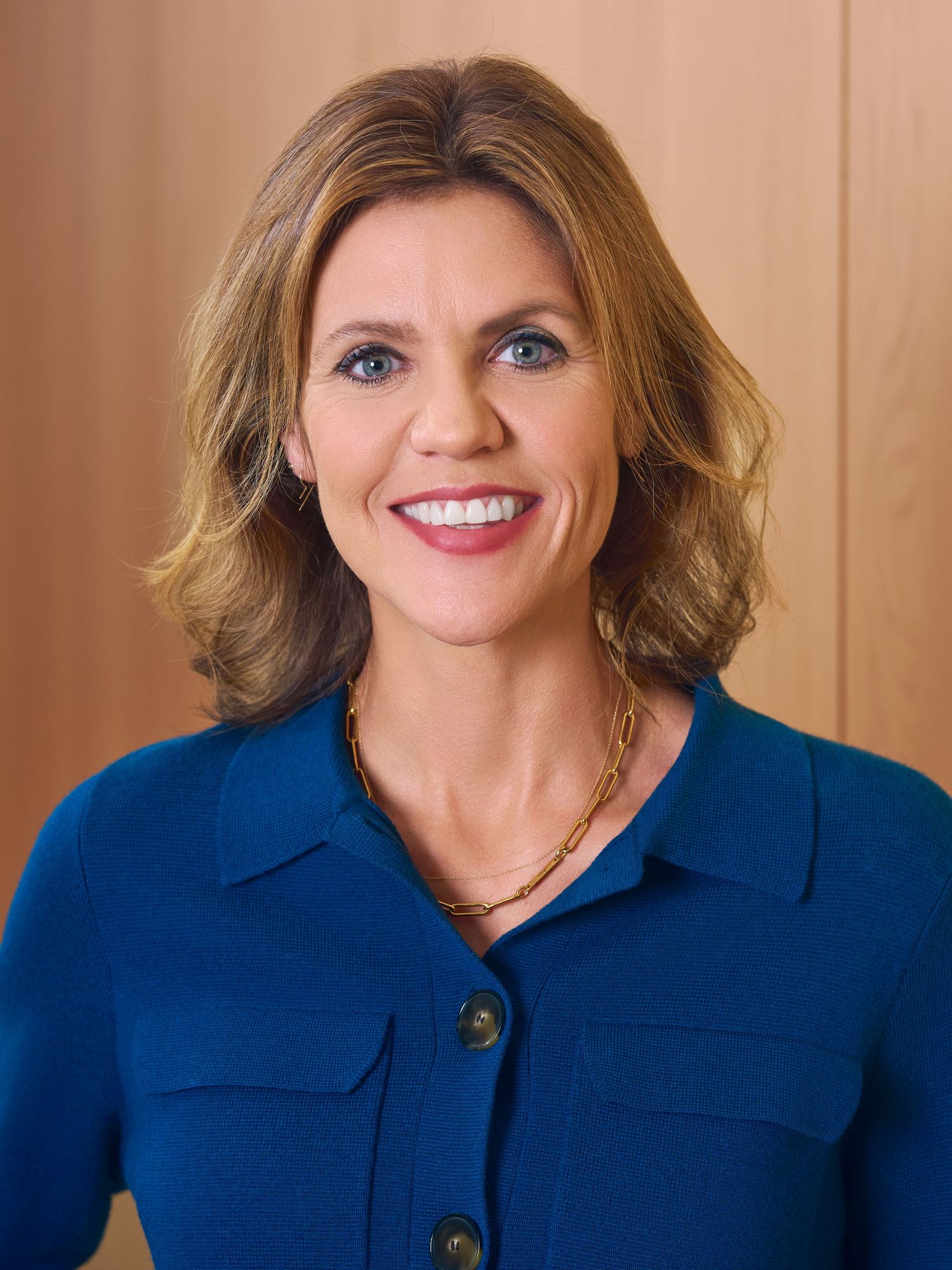 Professional headshot of a smiling woman with auburn hair wearing a blue blazer and gold necklace.