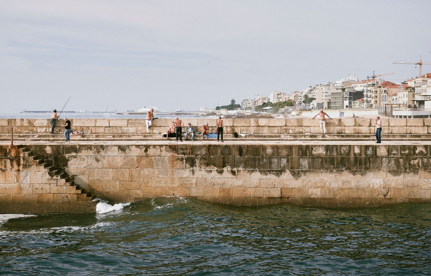Panoramic view of a stone harbor wall with people walking along the edge while waves crash against the structure.